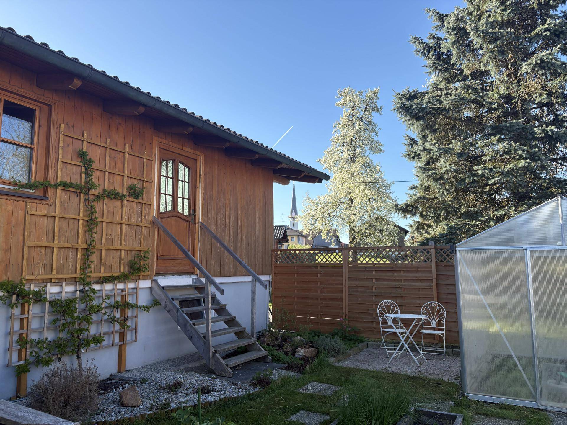 Kleiner Gartenbereich mit Holzfassade, Gewächshaus und Sitzgelegenheit.