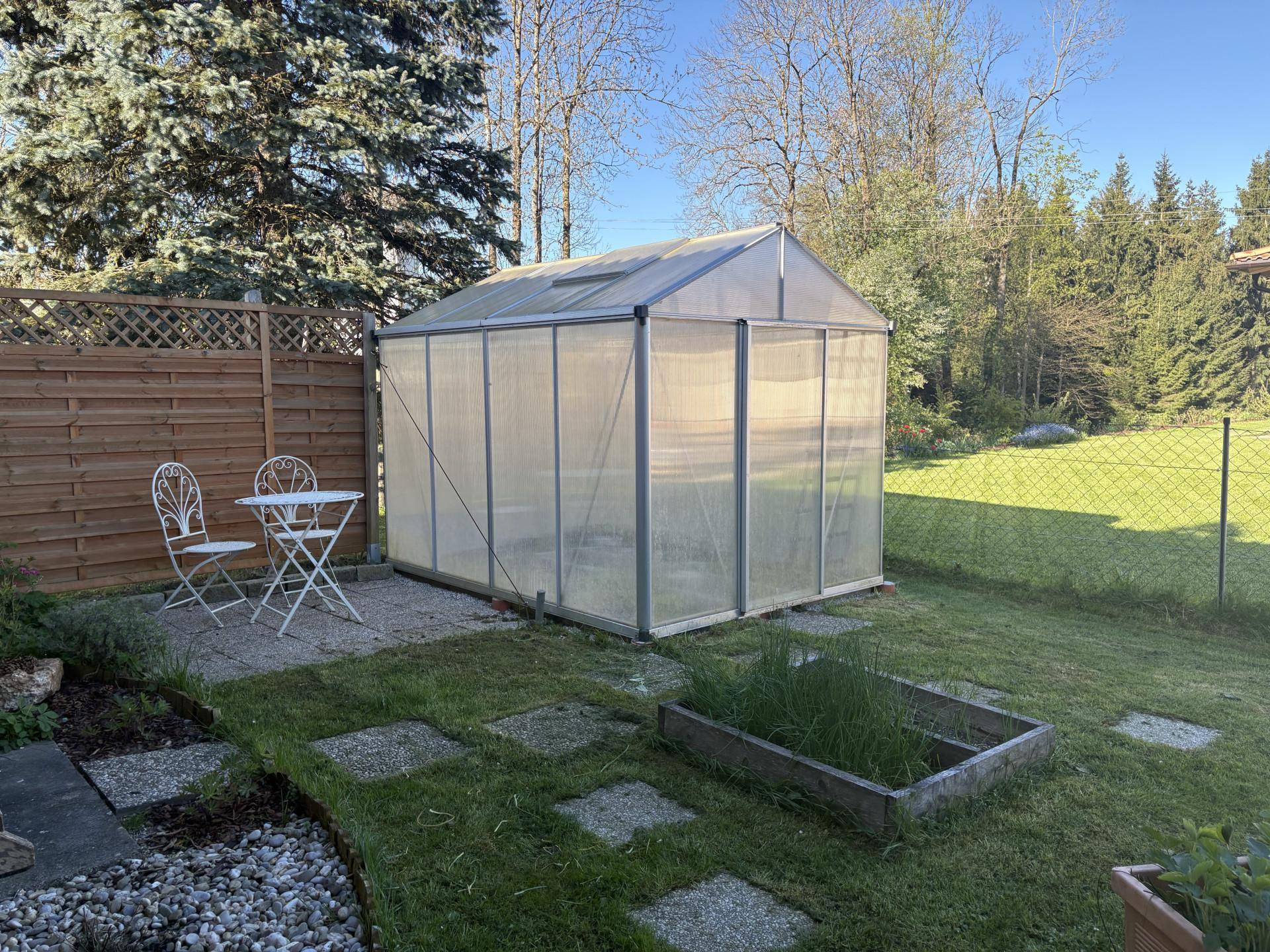 Sonniger Garten mit Gewächshaus, Hochbeet und Rasenfläche.
