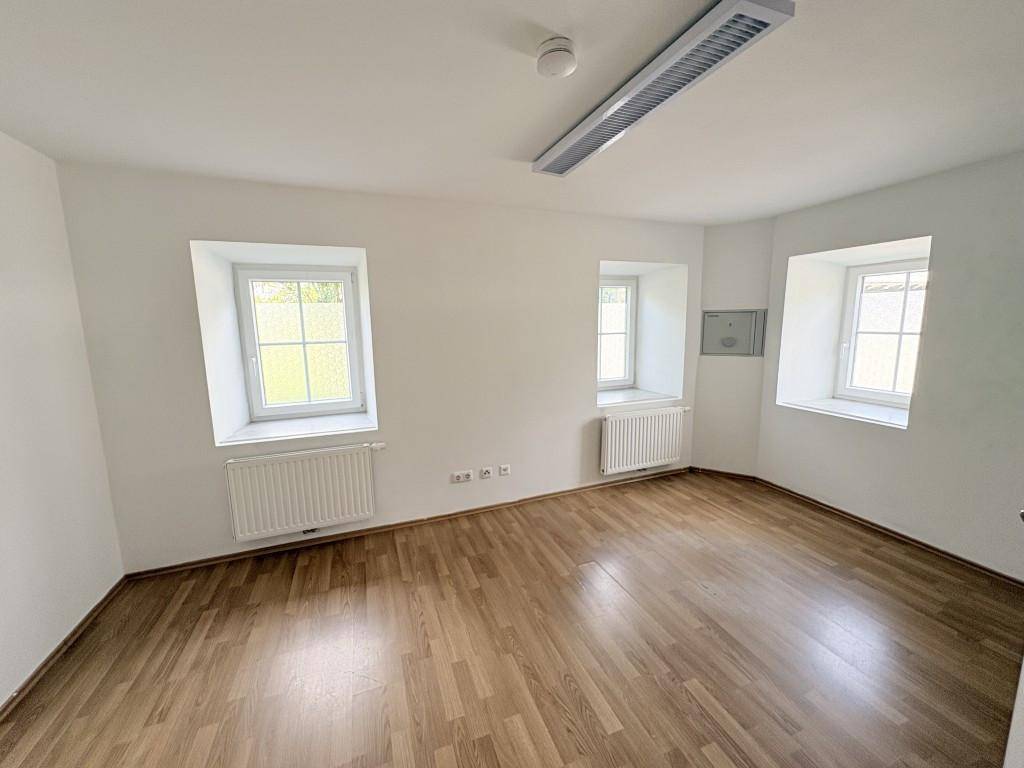 Heller, leerer Raum mit Holzboden, weißen Wänden und drei Fenstern.