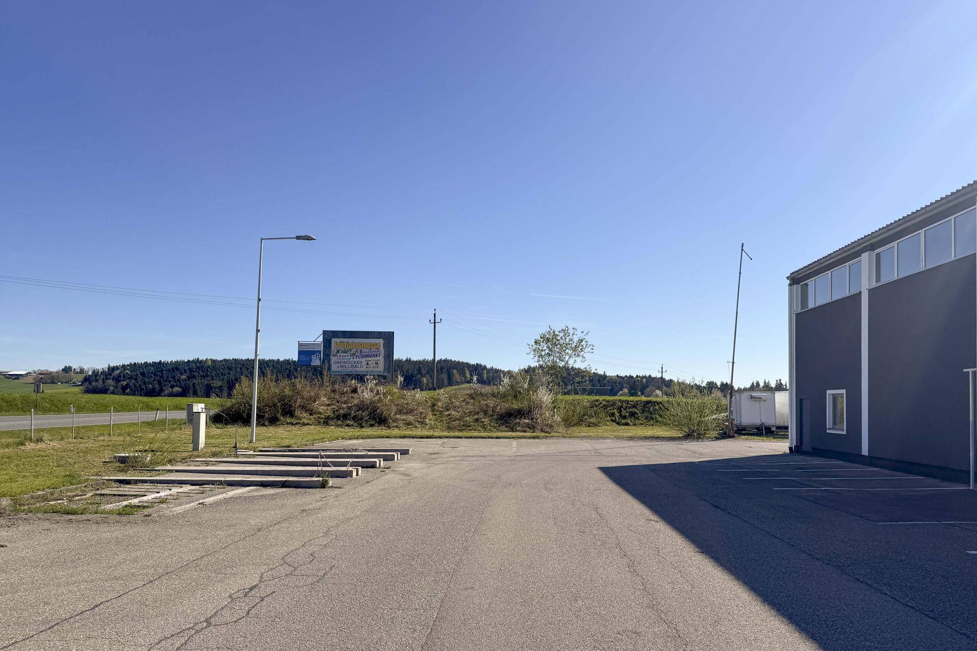 Parkplatz vor einem modernen Gewerbegebäude mit einem Werbeschild und Bäumen im Hintergrund unter blauem Himmel.