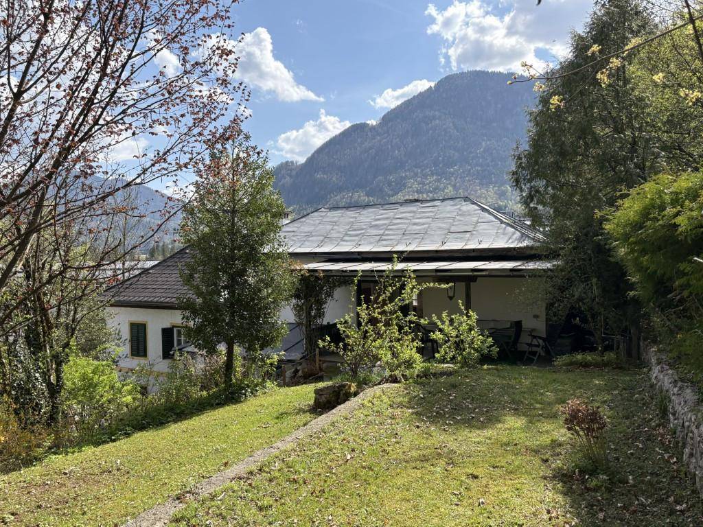 Traditionelles Wohnhaus mit Satteldach, umgeben von einem grünen Garten und Bäumen vor einer Bergkulisse.