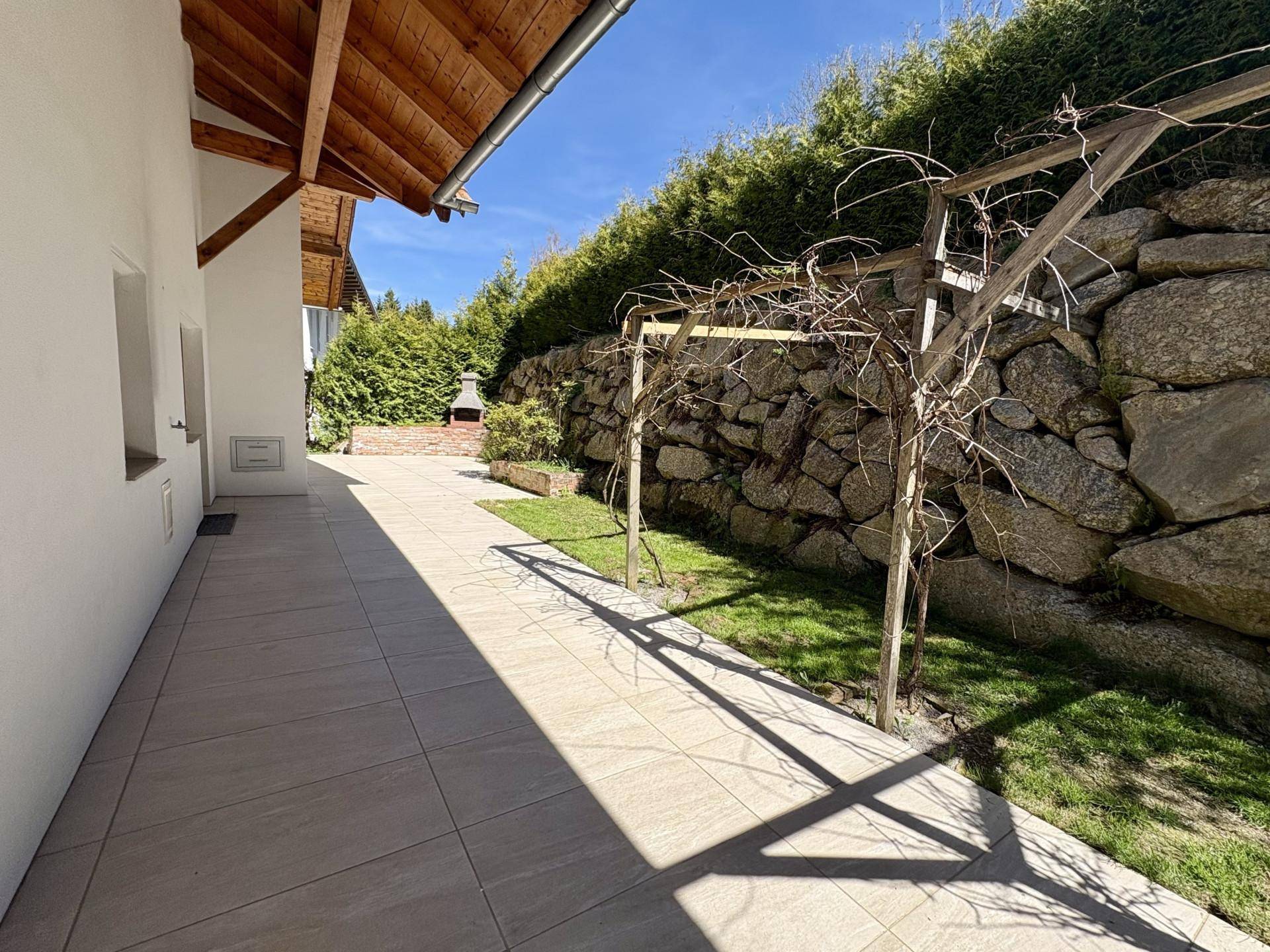 Sonniger Terrassenbereich mit Steinmauer, Grillplatz und Zugang zum Garten.