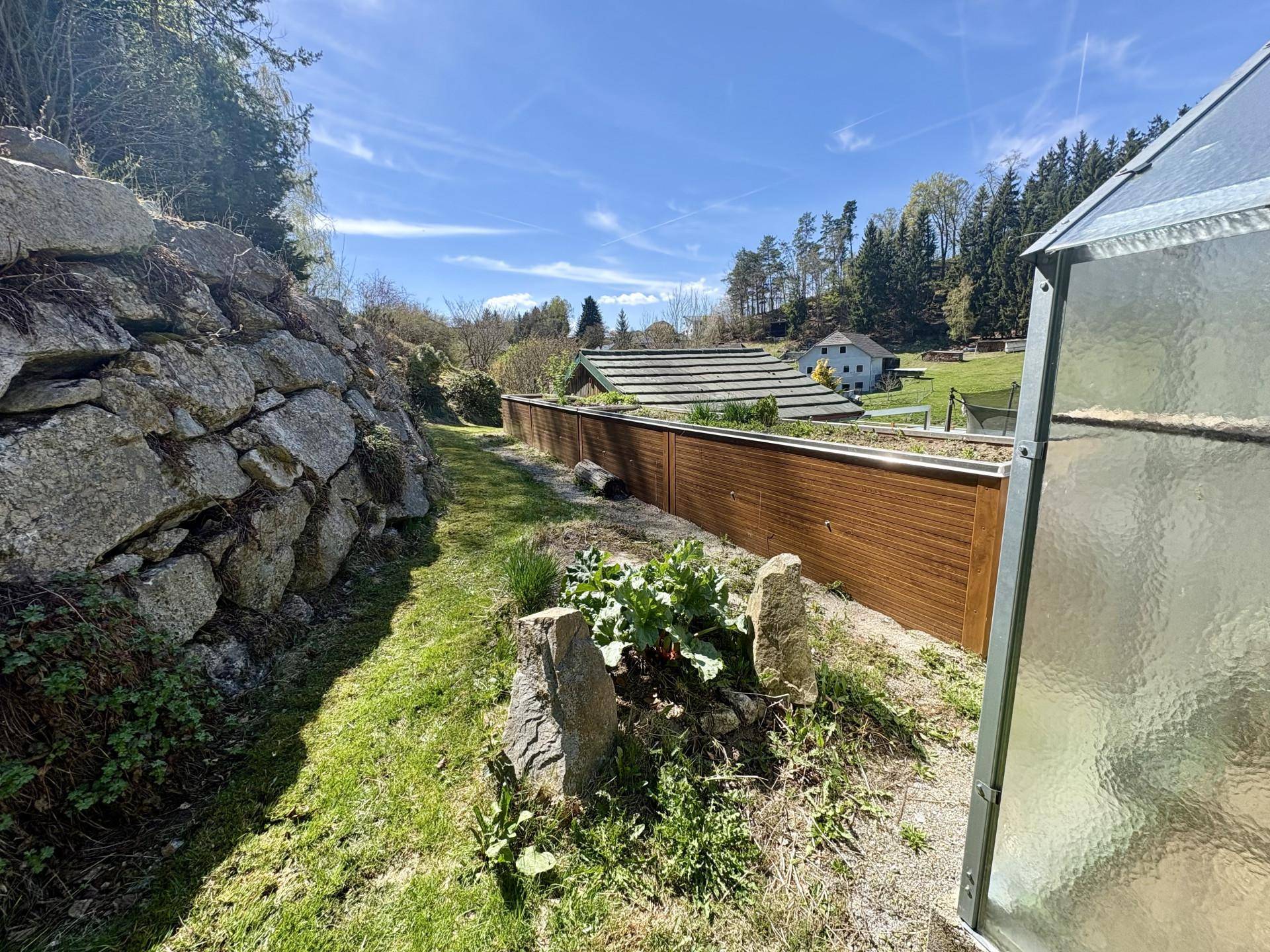 Gartenweg entlang einer Steinmauer mit Hochbeeten und Blick auf die umliegende Landschaft.