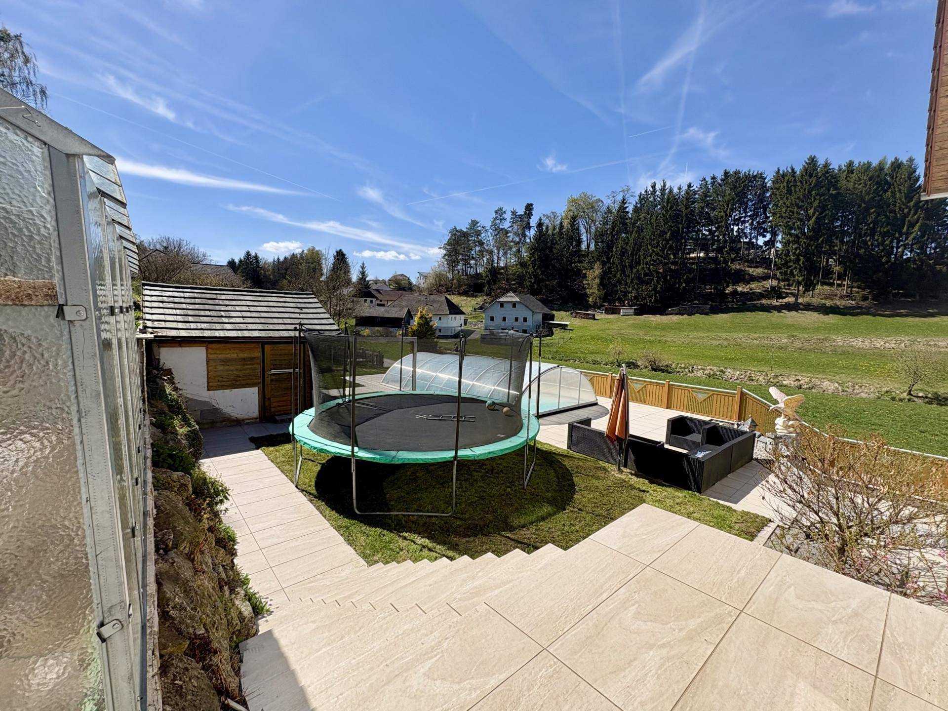 Großer Gartenbereich mit Trampolin, Sitzgelegenheiten und Blick auf den überdachten Pool.