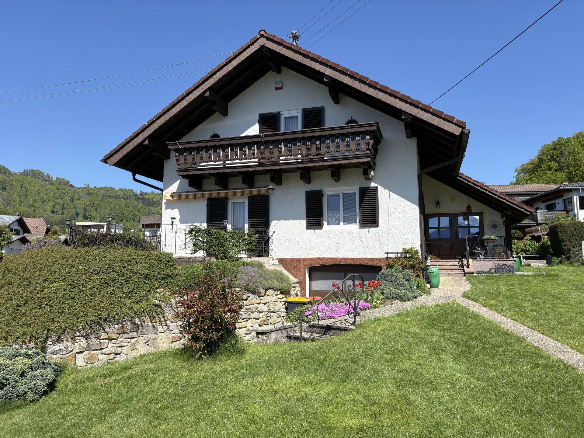 Traditionelles Haus mit Holzbalkon, grünen Fensterläden und gepflegtem Gartenbereich an einem sonnigen Tag.
