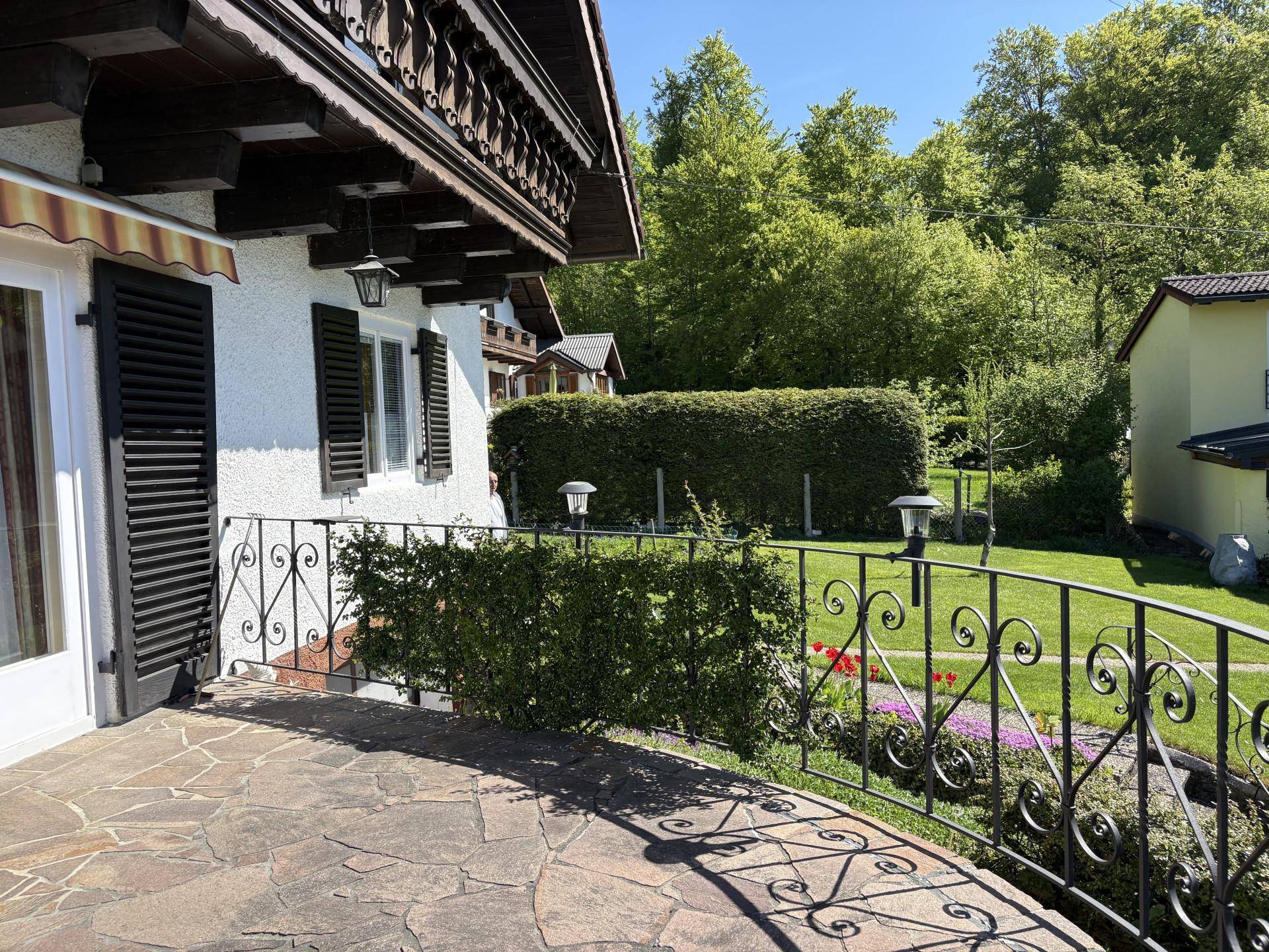 Sonnige Steinterrasse mit schmiedeeisernem Geländer und Blick auf den gepflegten Garten mit Hecke.