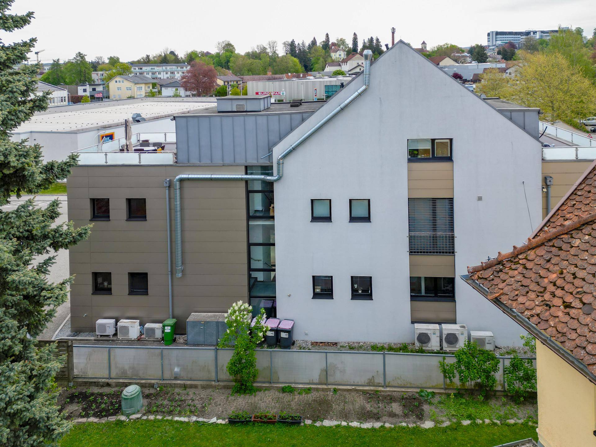 Rückansicht des Gebäudes mit sichtbaren Klimaanlagen und technischen Installationen, umgeben von Bäumen.