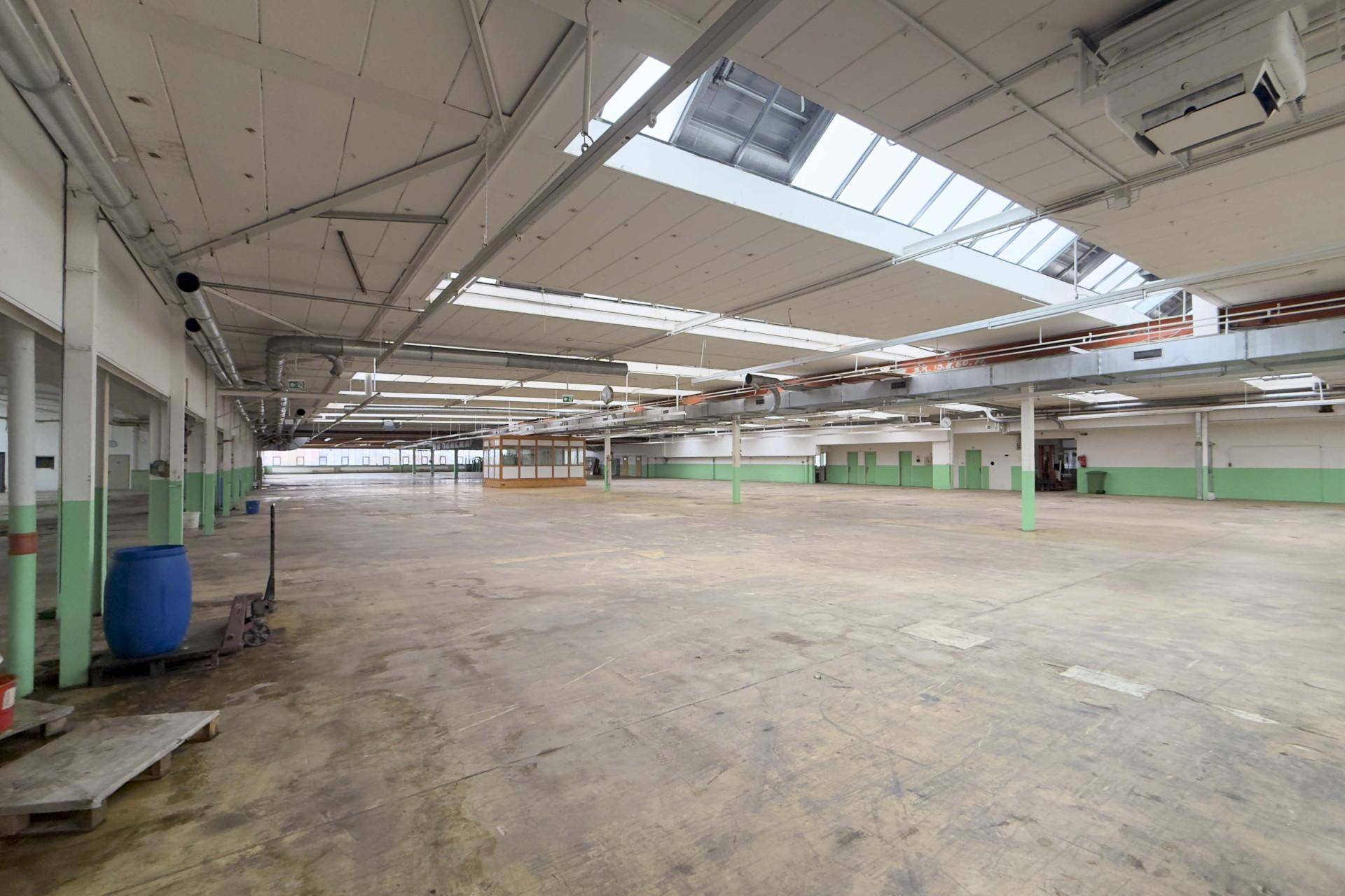 Weitläufige Industriehalle mit Betonboden, Oberlichtern und sichtbaren Lüftungsrohren, bereit zur flexiblen Nutzung.