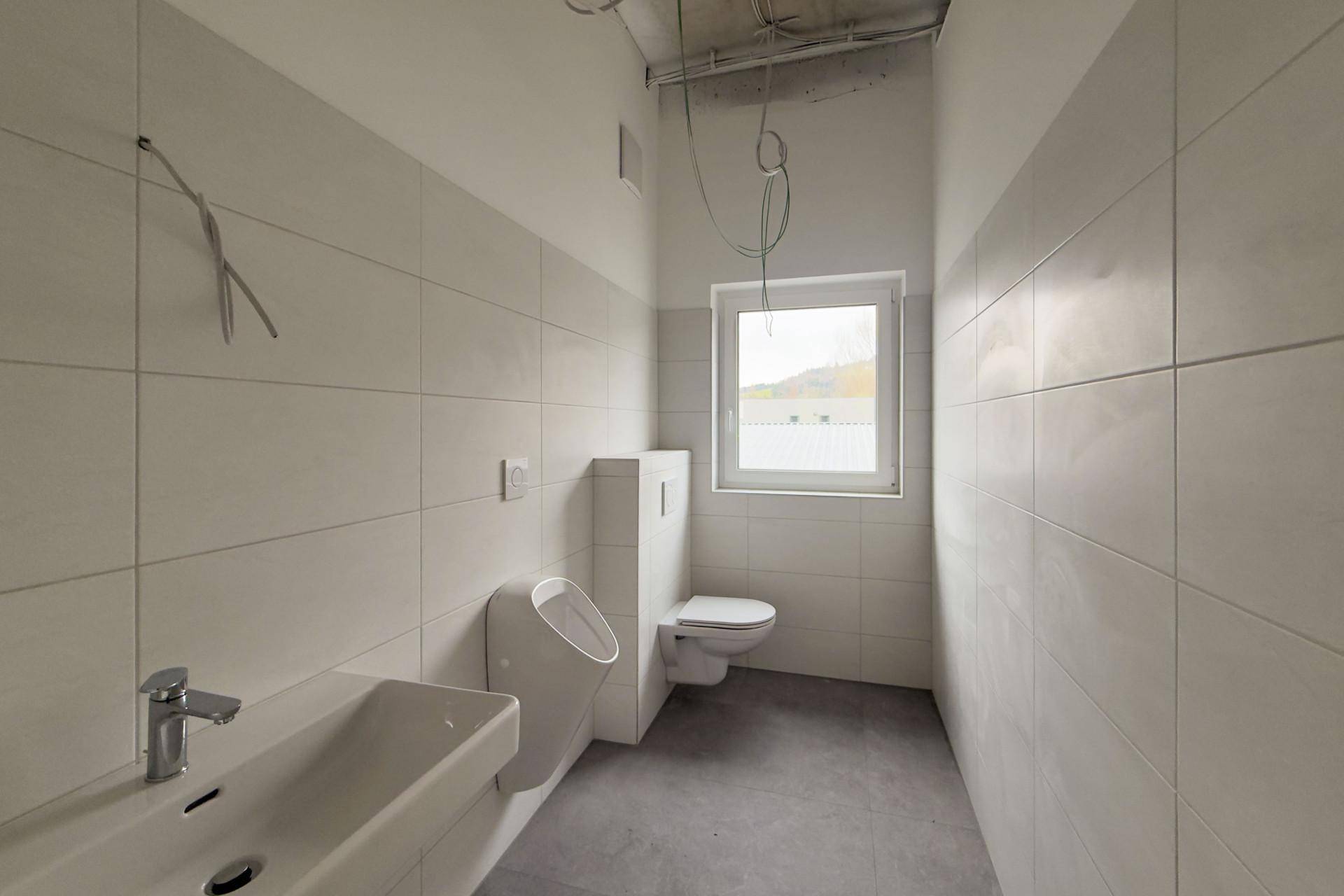 Gefliestes Badezimmer mit Waschbecken, Toilette, Urinal und Fenster, noch unfertig.