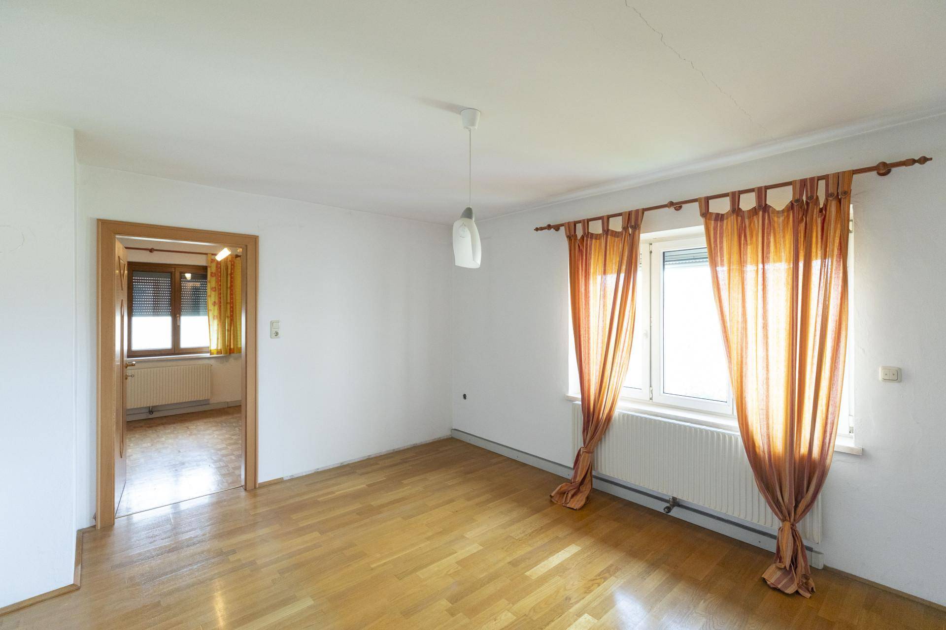 Helles Zimmer mit Holzboden, weißen Wänden und einem Fenster mit orangefarbenen Vorhängen.