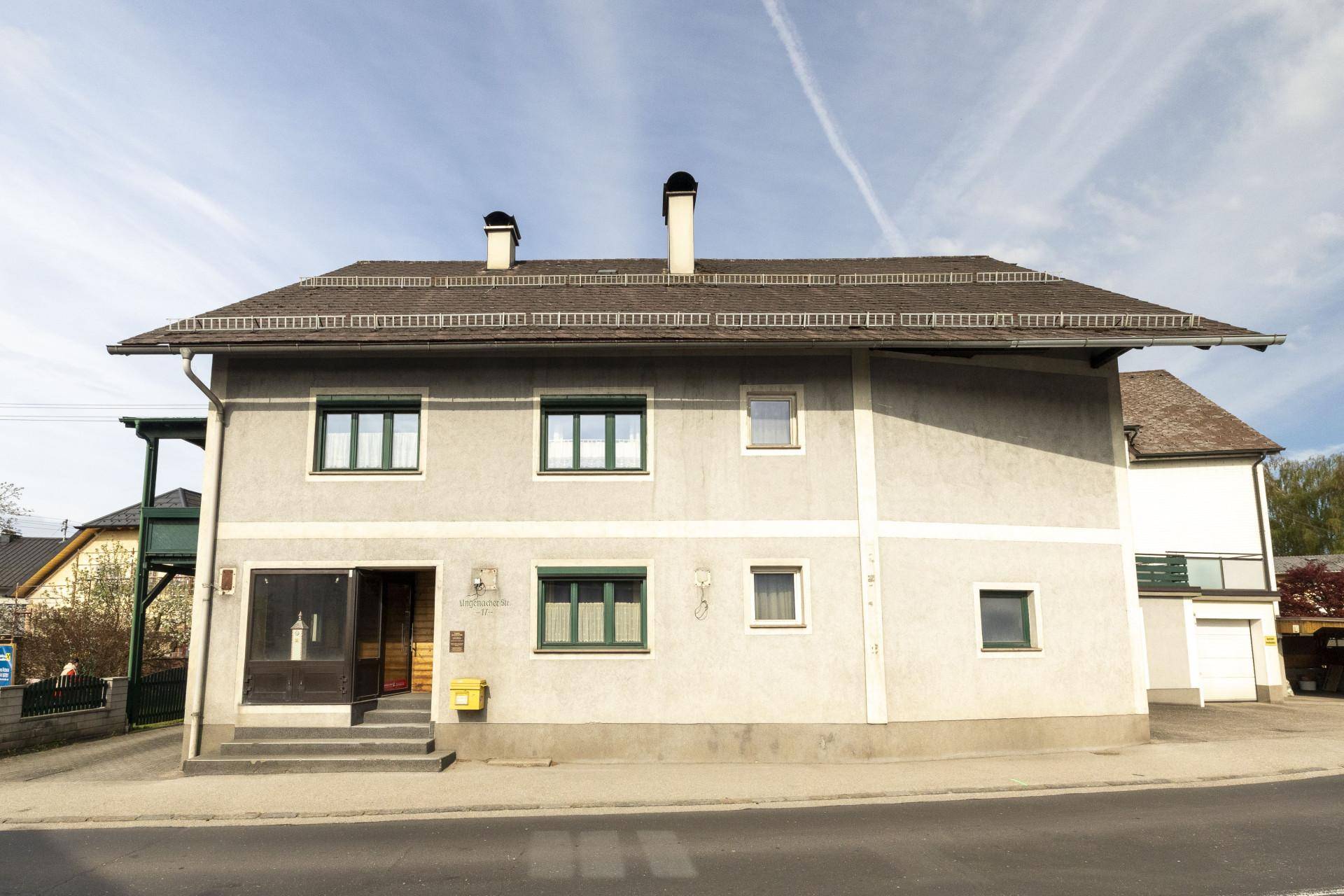 Seitenansicht eines grauen Hauses mit mehreren Fenstern und zwei Schornsteinen unter blauem Himmel.