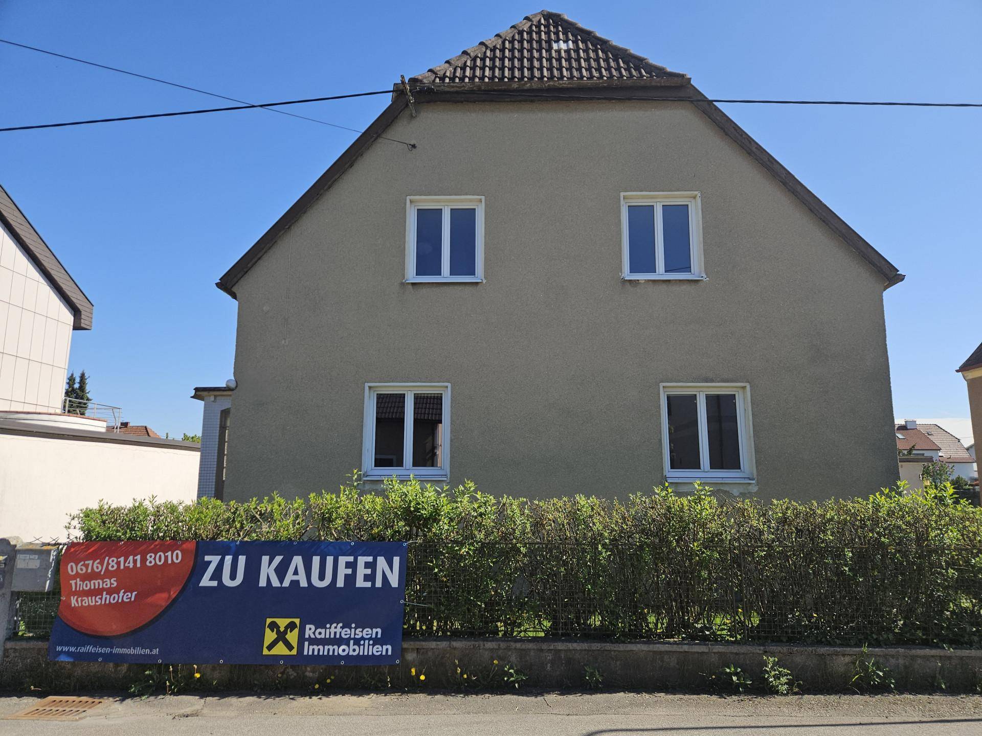 Vorderansicht des Hauses mit Satteldach und einem "Zu Kaufen" Schild im Vorgartenbereich.