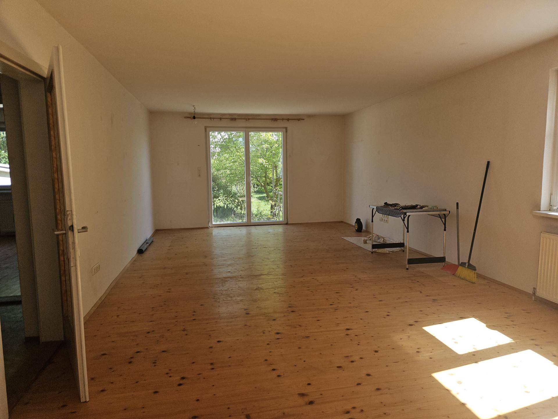 Geräumiges Wohnzimmer mit Holzdielenboden und großem Fenster mit Blick ins Grüne.