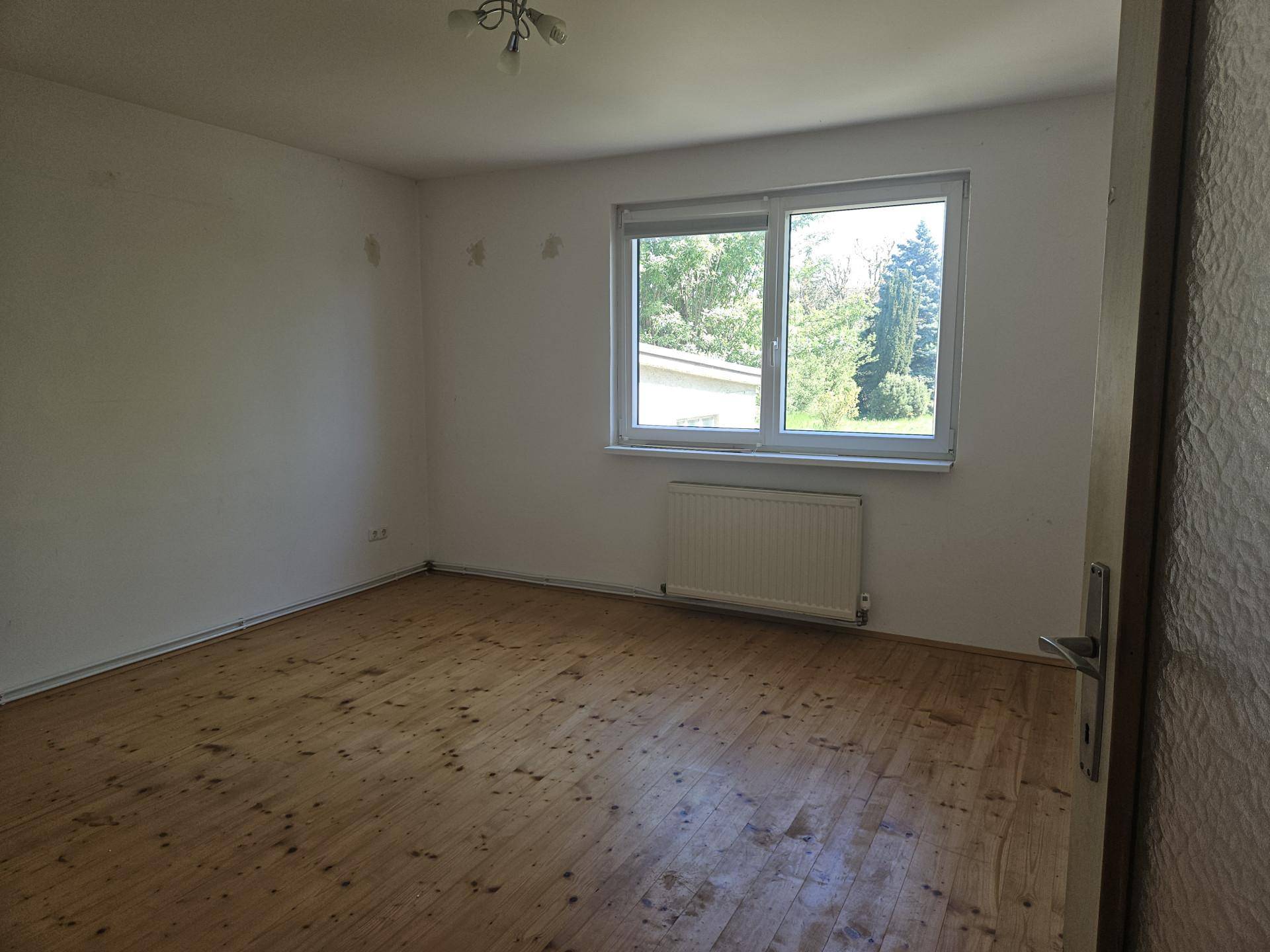 Helles Zimmer mit Holzdielenboden, Heizkörper und Fenster mit Blick auf Bäume.