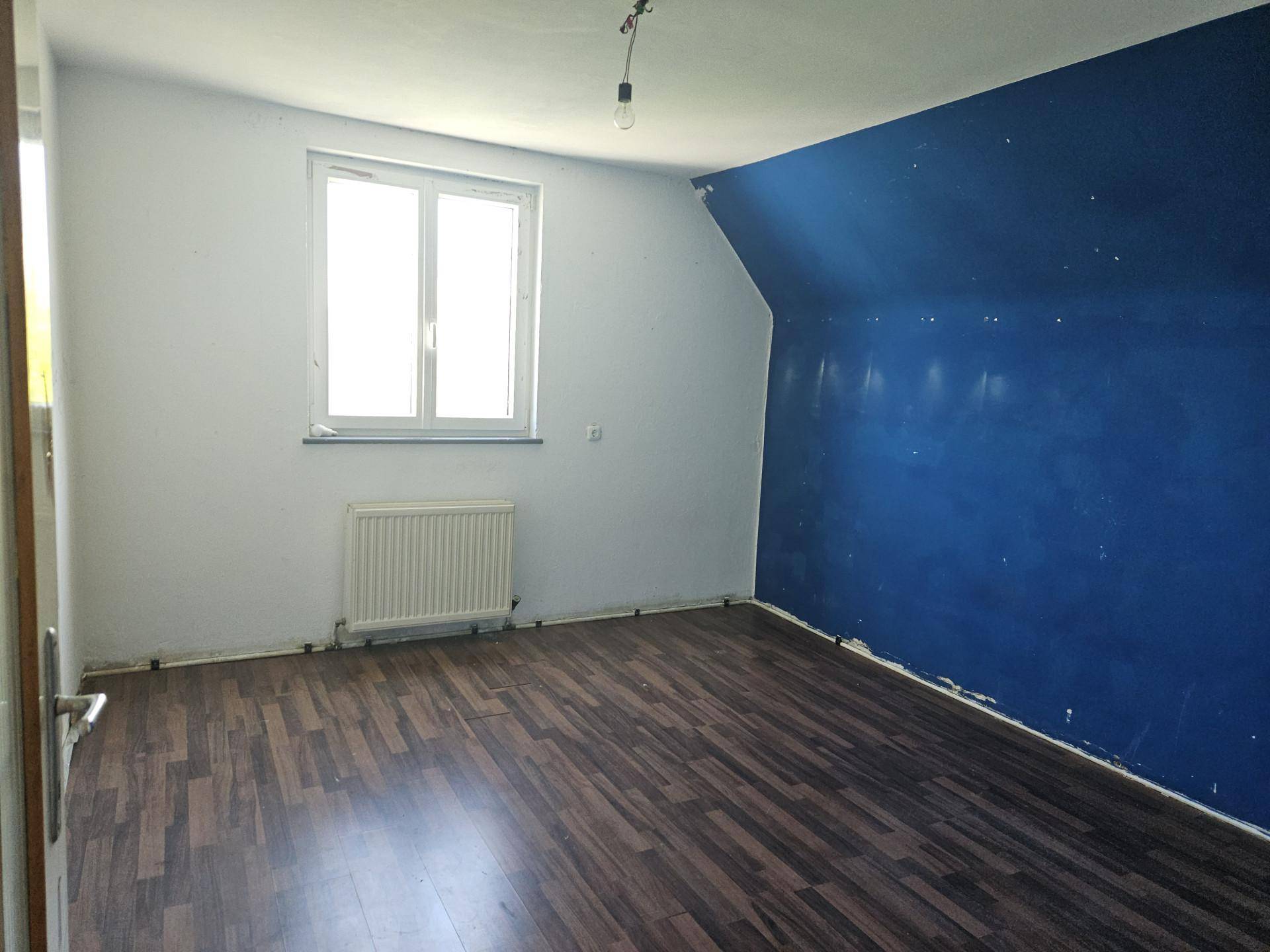 Zimmer mit dunkelblau gestrichener Wand, Laminatboden und Fenster im Dachgeschoss.