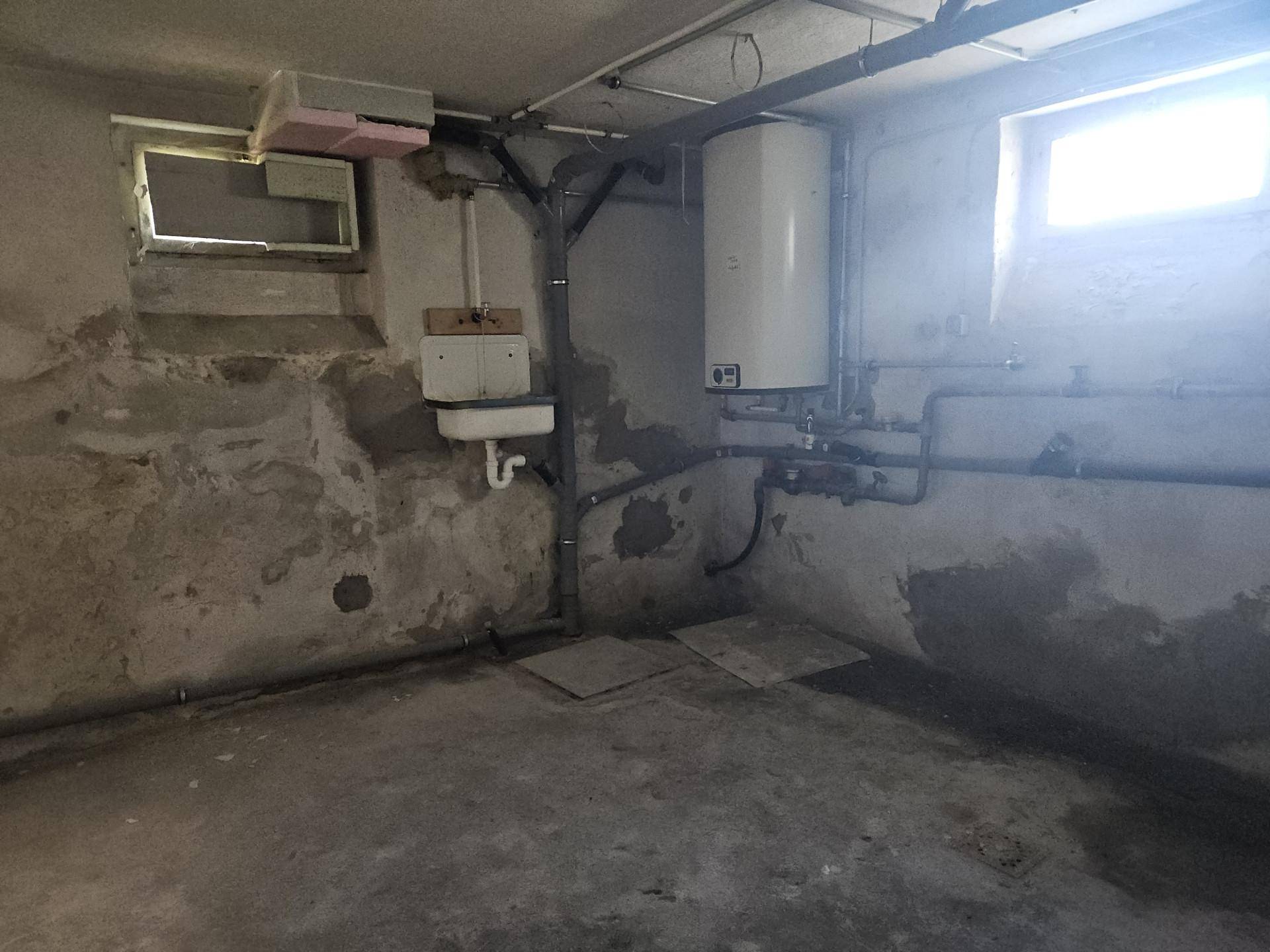 Kellerraum mit Waschbecken, Warmwasserboiler und sichtbaren Rohren, renovierungsbedürftig.
