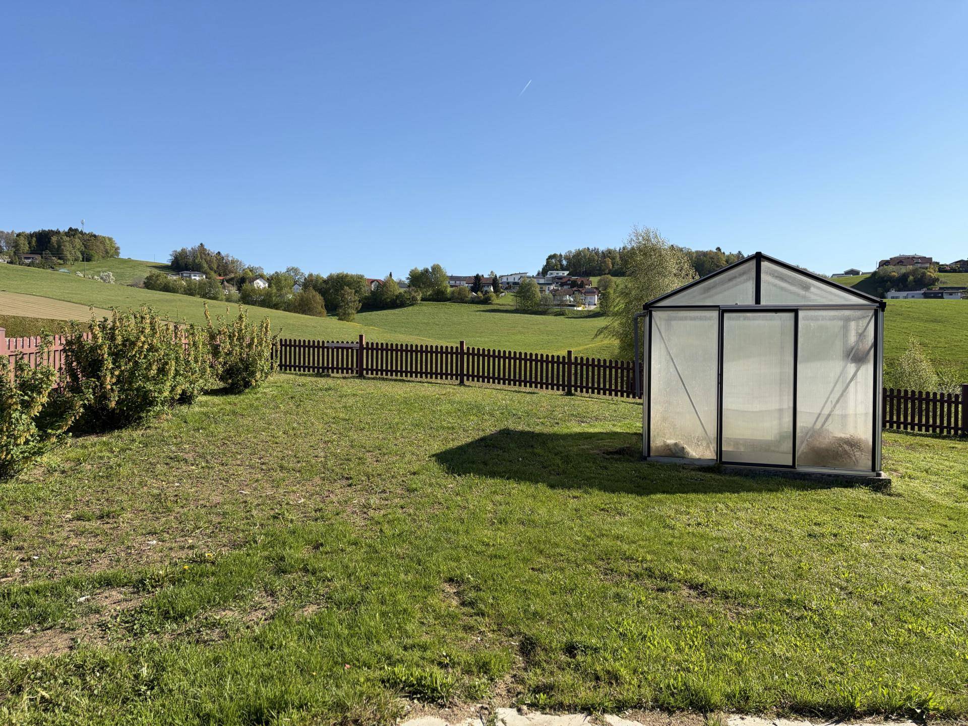Großer Garten mit Gewächshaus und Holzzaun, umgeben von sanften grünen Hügeln.