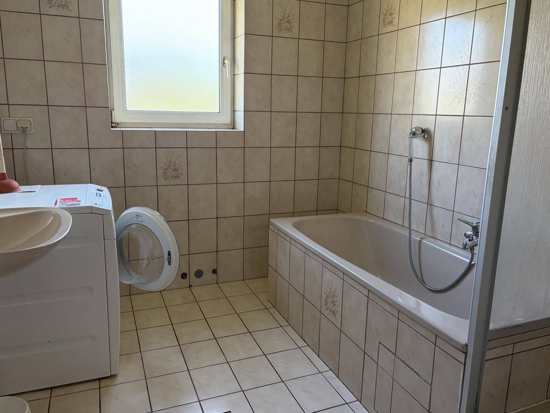 Badezimmer mit Badewanne, Waschmaschine und Fenster, gefliesten Wänden und Boden.