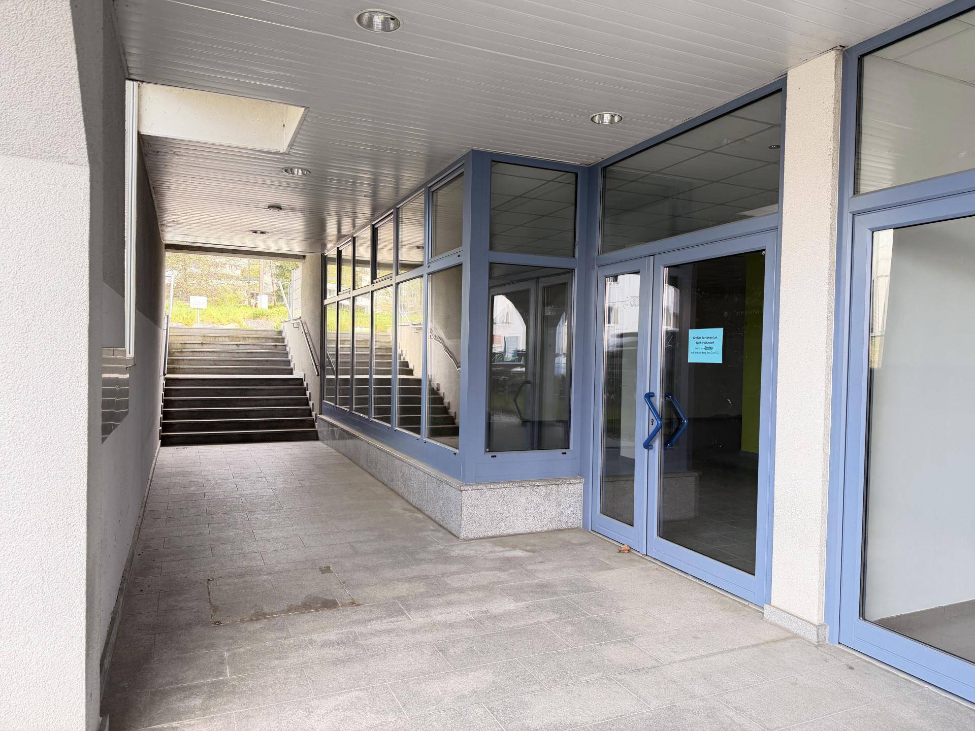 Überdachter Außenbereich mit Treppe und großen Glasfronten, die zu einem Eingangsbereich führen.