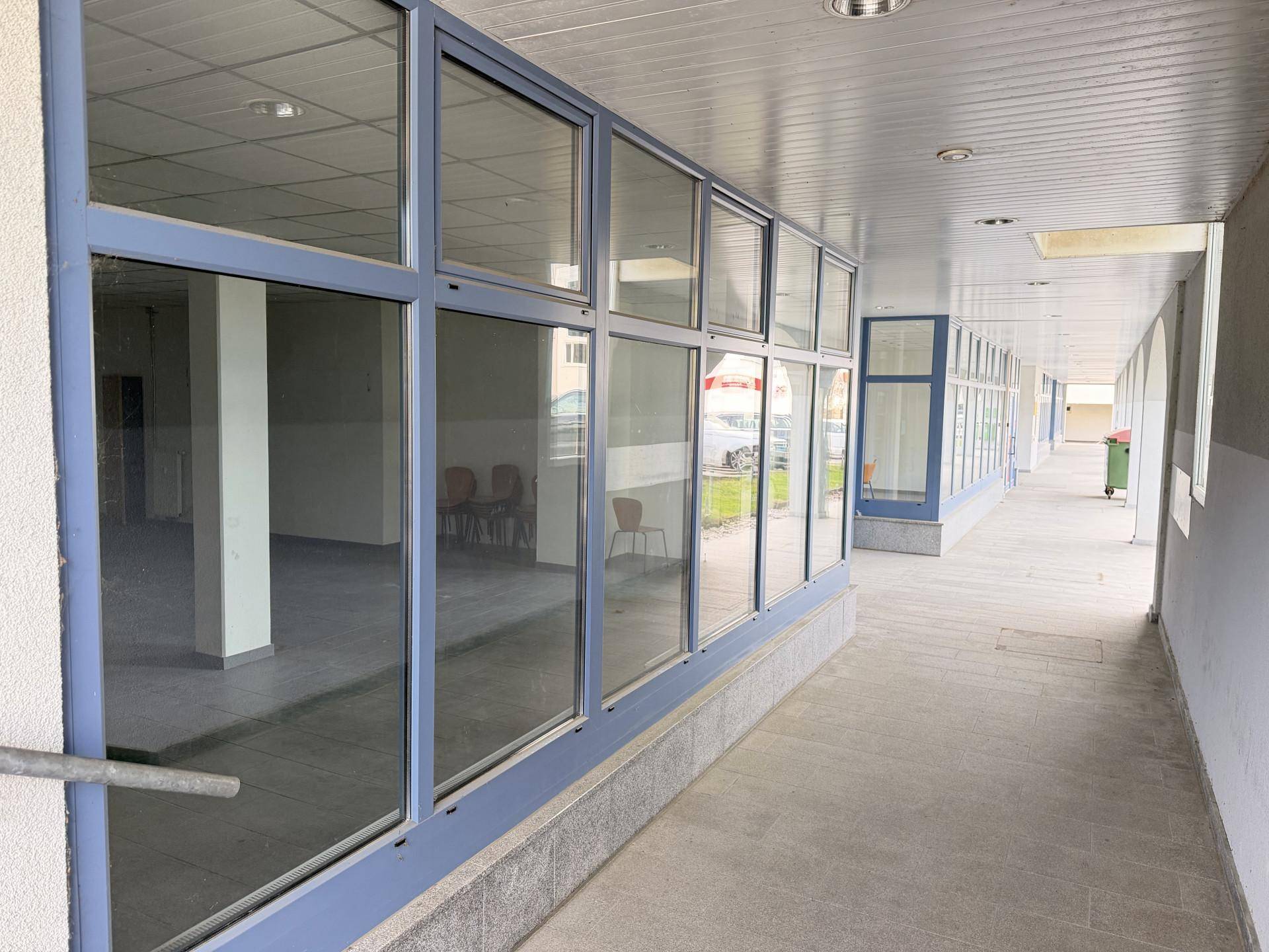 Langer überdachter Gang entlang der Gebäudefassade mit durchgehenden Fensterfronten, die Einblicke in die Innenräume gewähren.
