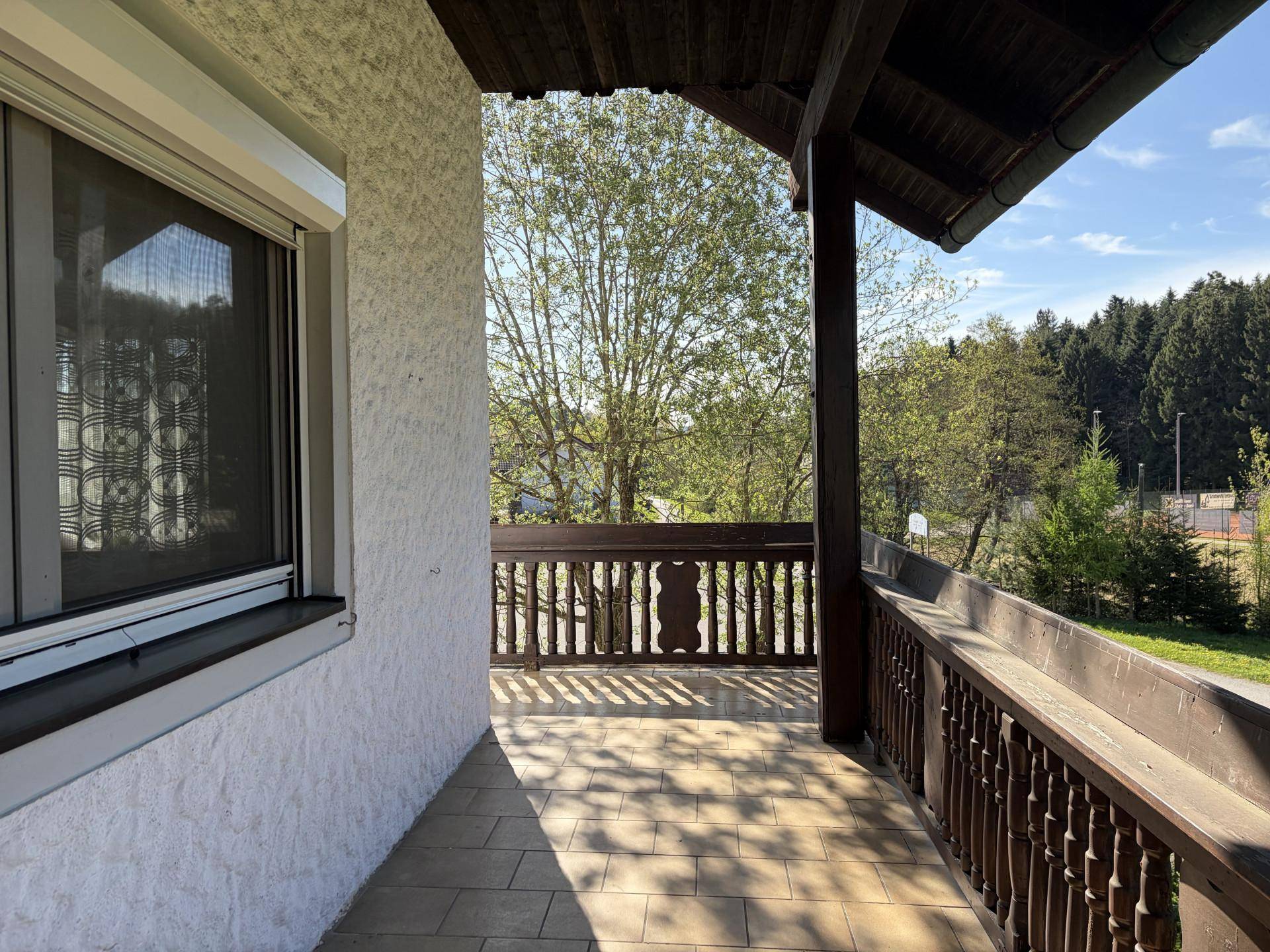Gedeckter Balkon mit Fliesenboden und traditionellem Holzgeländer, bietet Blick ins Grüne.