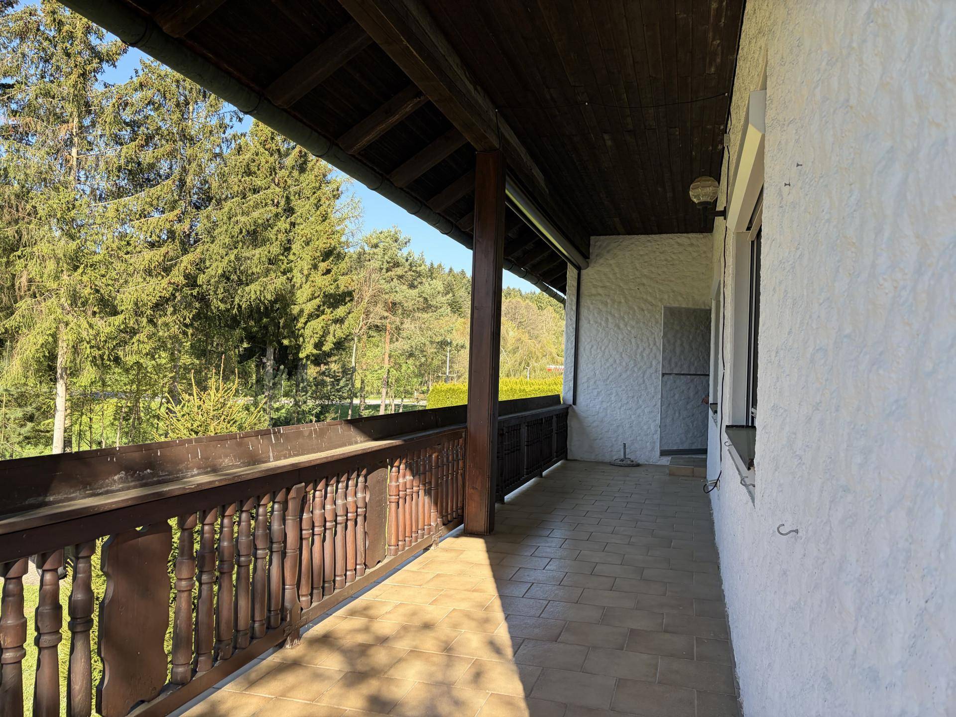 Langer, überdachter Balkon mit Fliesenboden und Holzbalken, umgeben von hohen Bäumen.