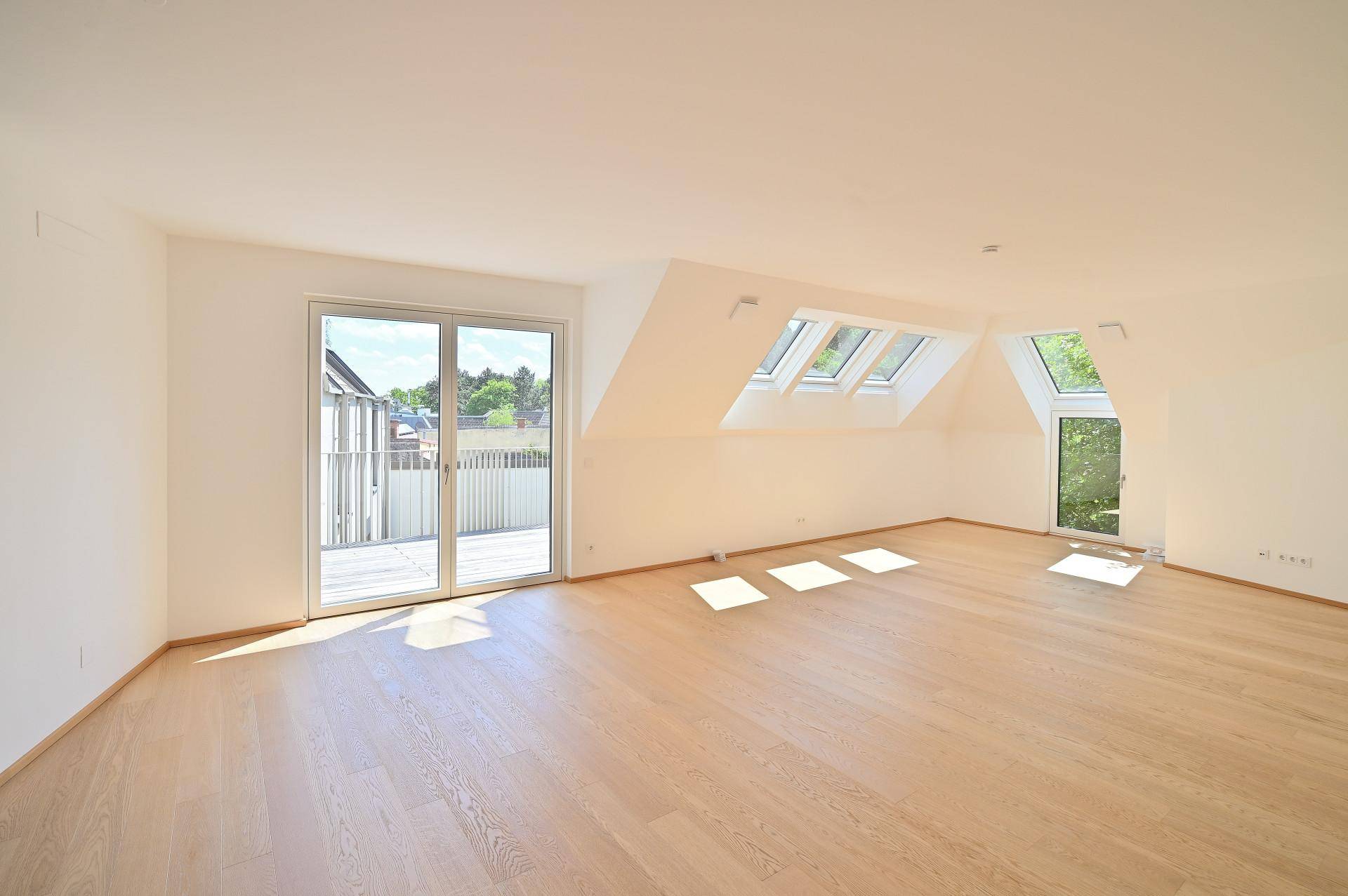 Geräumiges, leeres Wohnzimmer mit Holzboden, Dachfenstern und Zugang zum Balkon.