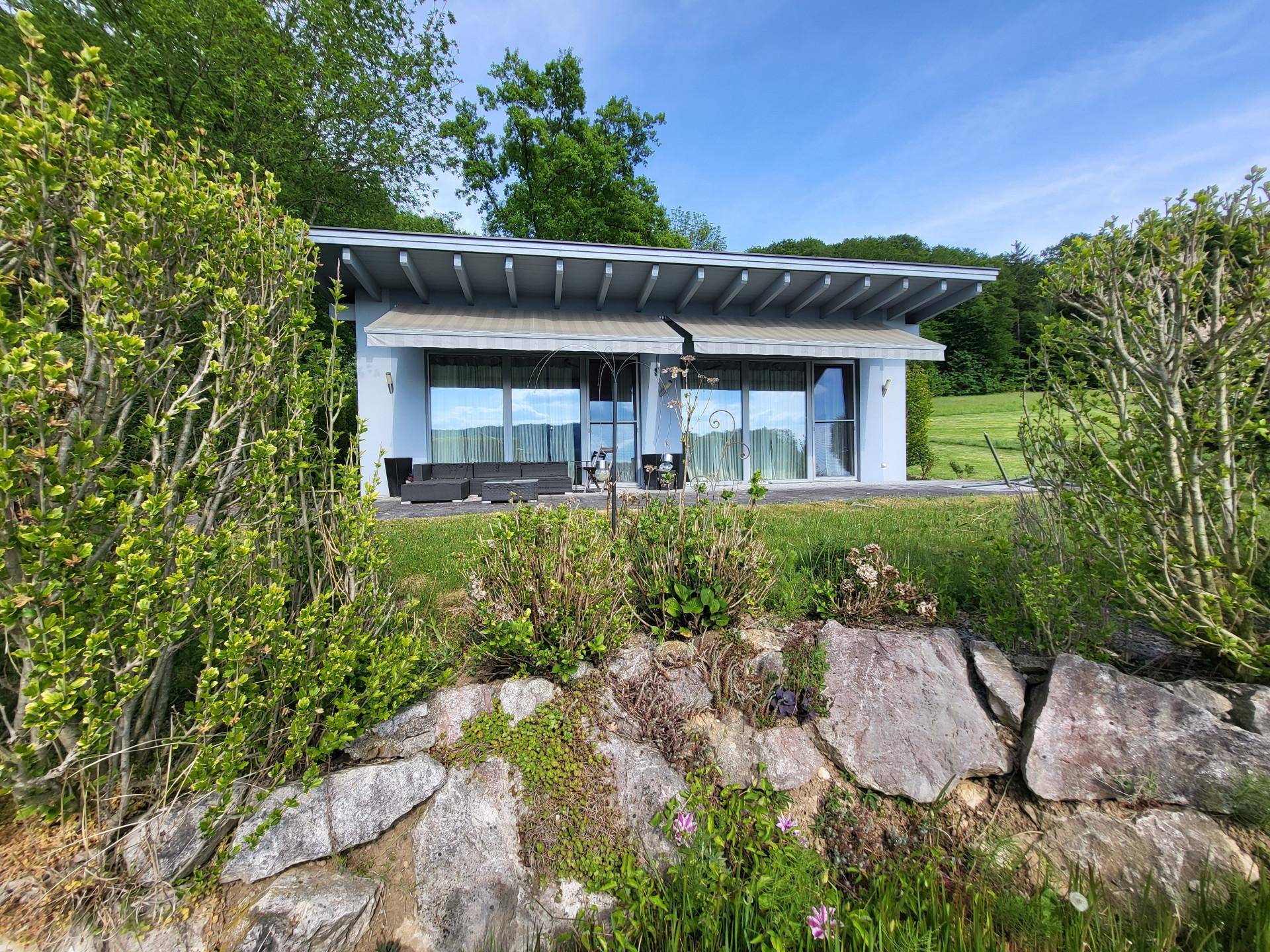 Helles Haus mit großer Terrasse und Sitzgelegenheiten, eingebettet in einen grünen Garten.