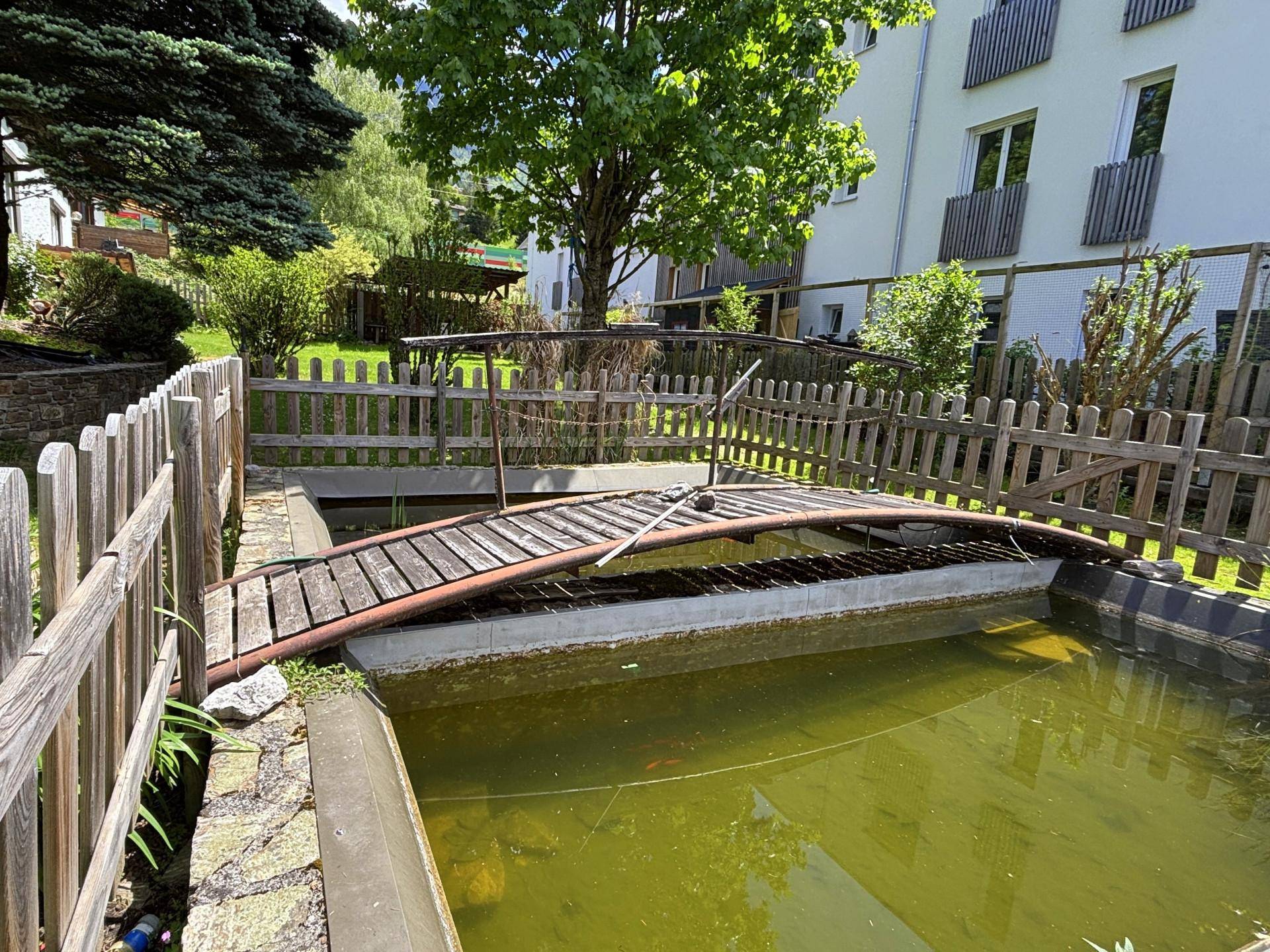 Gartenteich mit kleiner Holzbrücke und umzäuntem Bereich im Grünen.