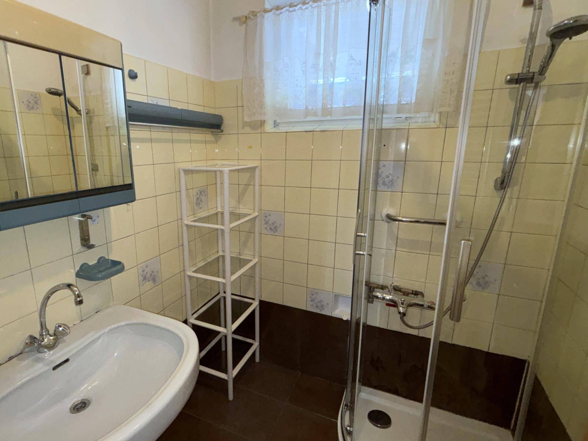 Badezimmer mit Dusche, Waschbecken und hellen Fliesen im 80er-Jahre-Stil.