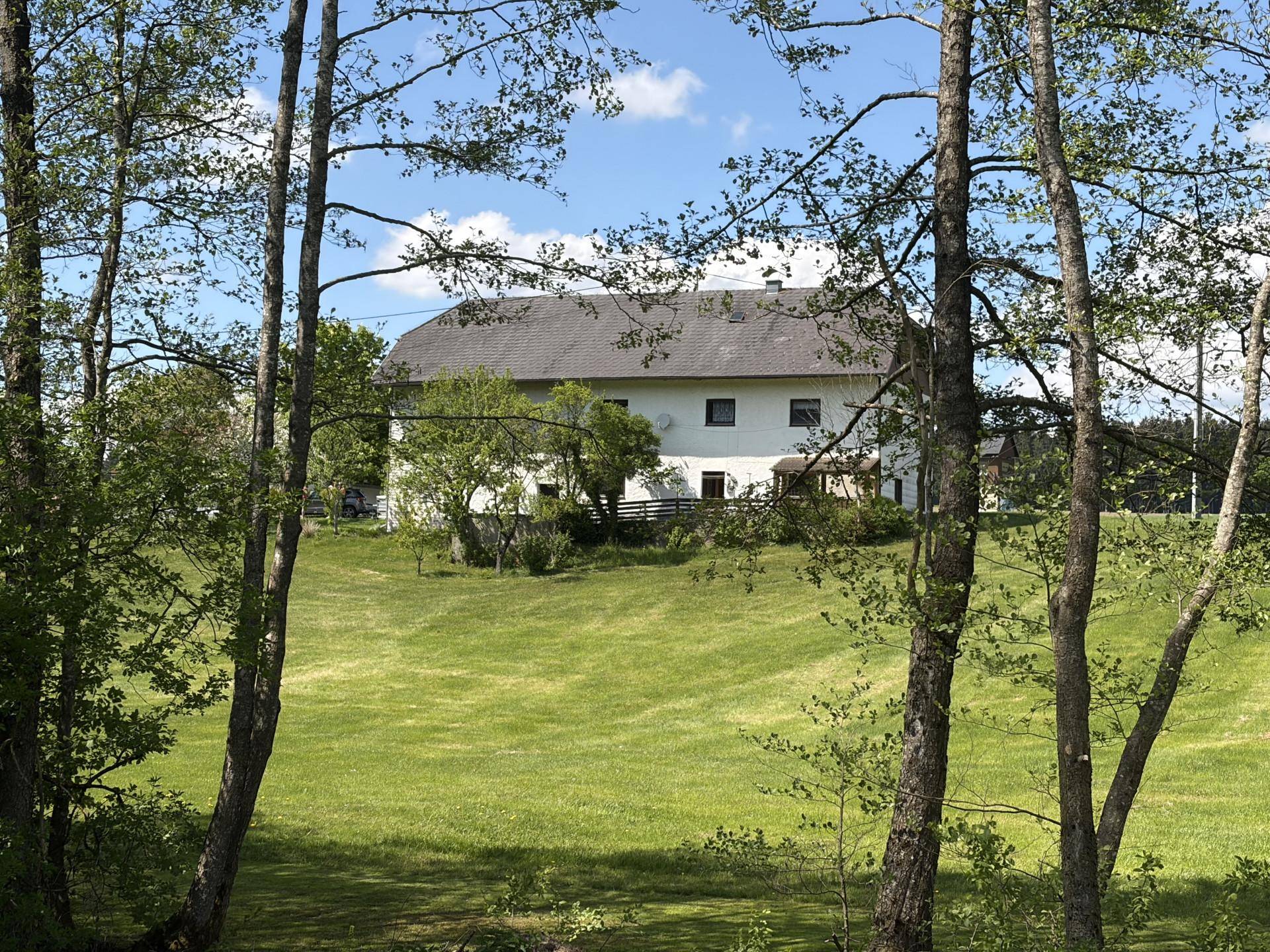 Ländliches Wohnhaus, umgeben von grünen Wiesen und Bäumen, unter einem strahlend blauen Himmel.