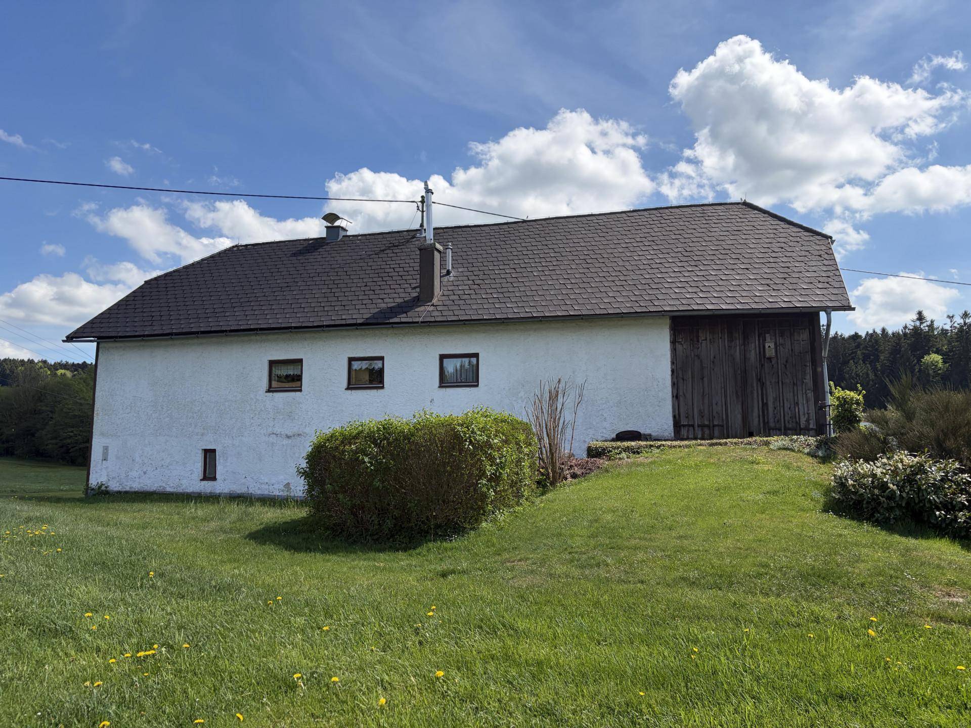 Rückansicht eines ländlichen Hauses mit weißer Fassade, Holztür und dunklem Dach, umgeben von Wiesen.