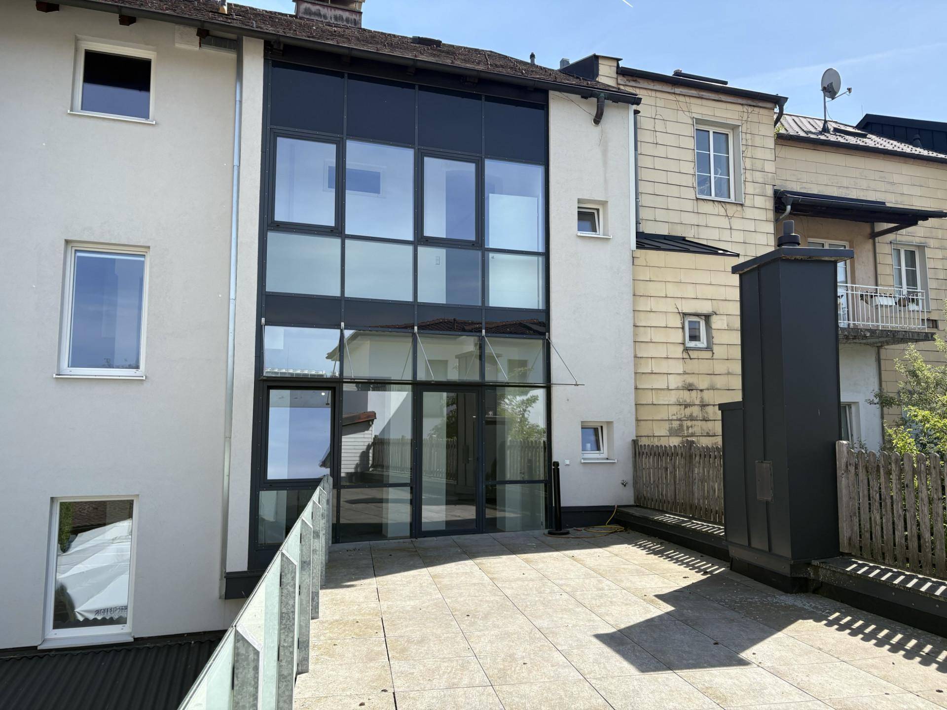 Moderne Gebäudefassade mit großer Glasfront und einer geräumigen, gepflasterten Terrasse im Freien.