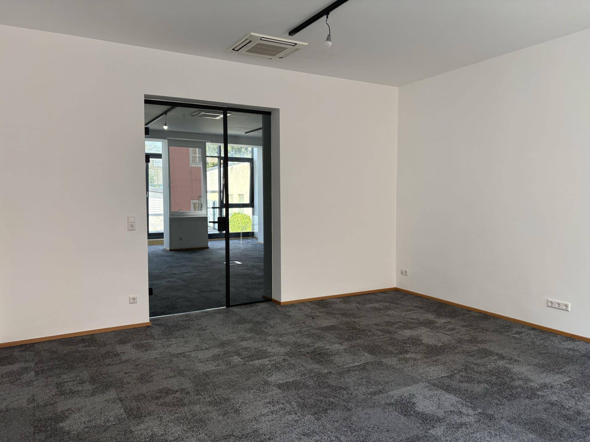 Heller Büroraum mit grauem Teppichboden, weiß gestrichenen Wänden und einer modernen Glasschiebetür.
