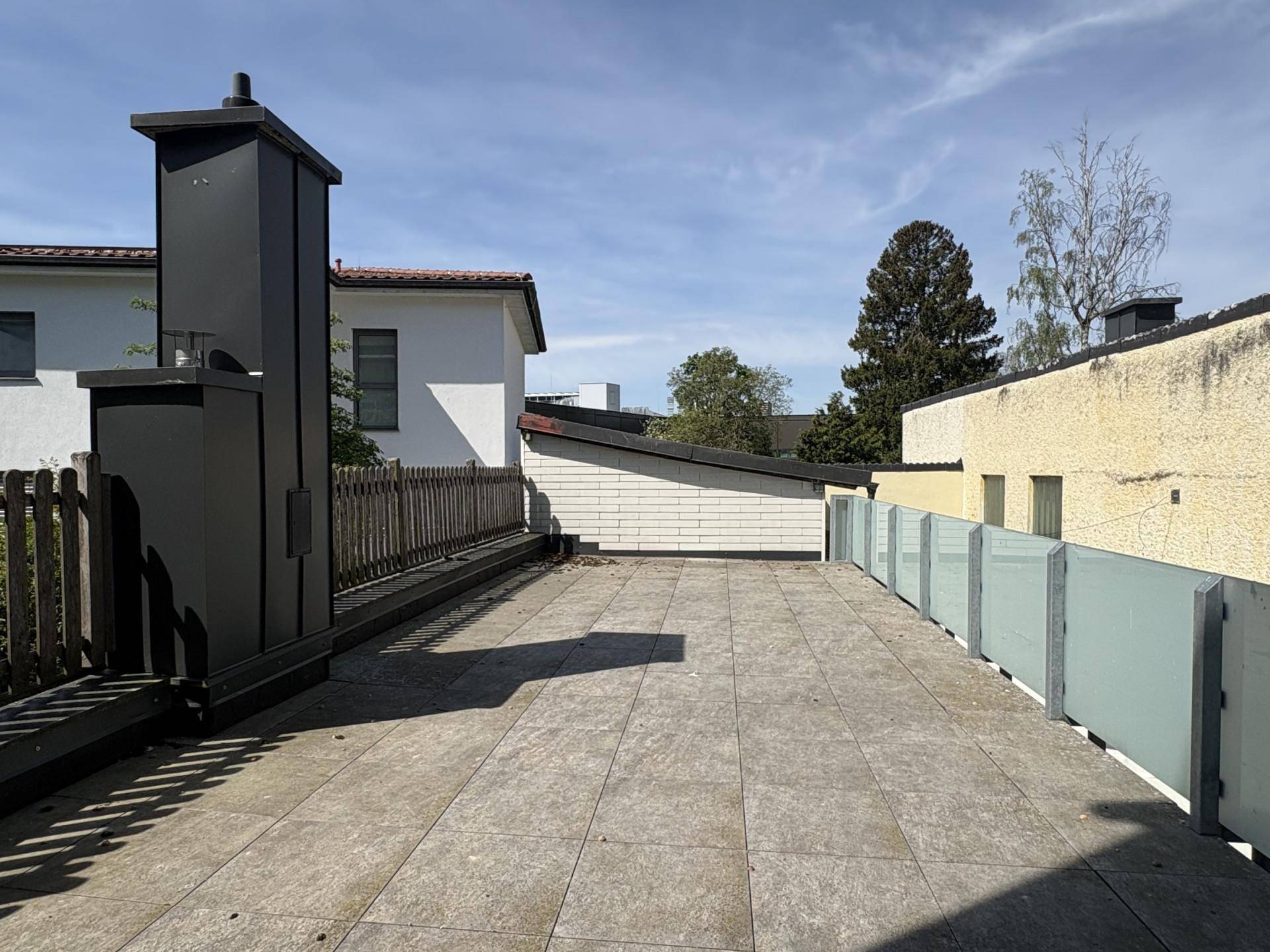 Weitläufige, gepflasterte Dachterrasse mit Glasgeländer und Blick auf die umliegenden Gebäude und Bäume.