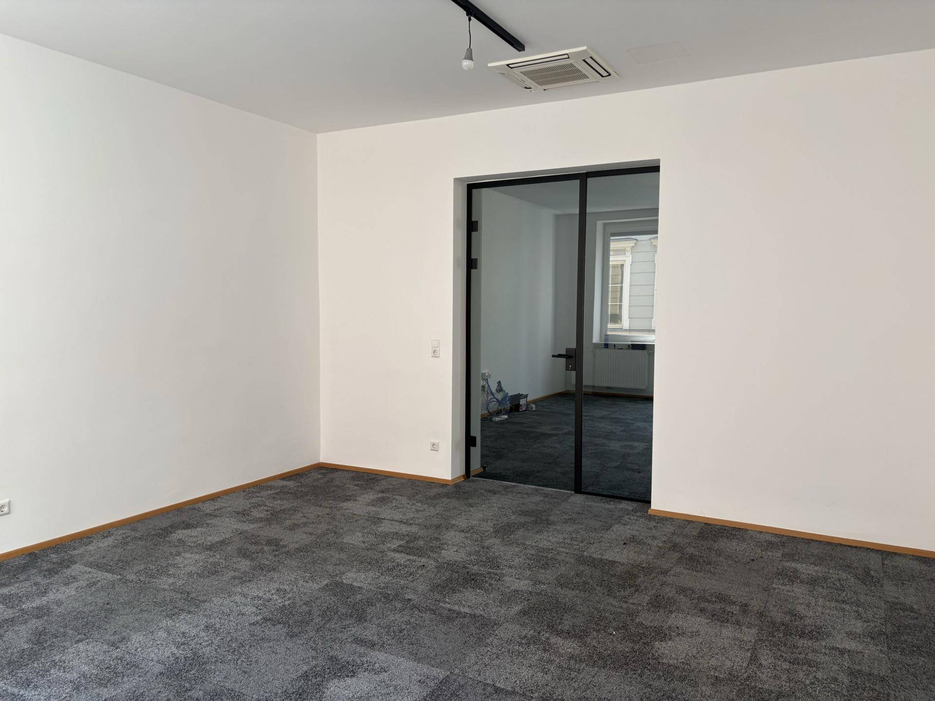 Geräumiger Büroraum mit grauem Teppichboden, weiß gestrichenen Wänden und einer modernen Glastür.