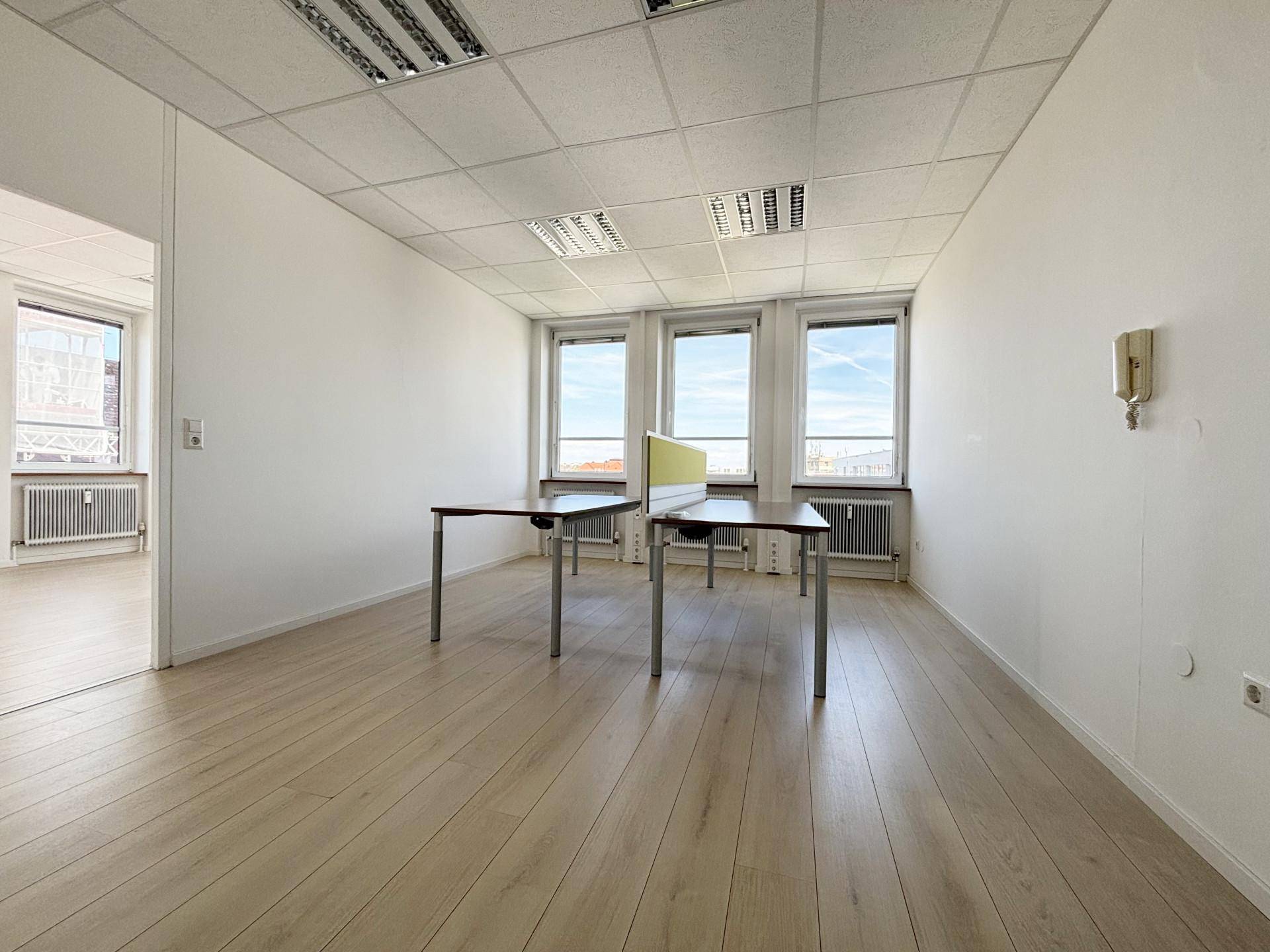 Bürofläche mit hellem Laminatboden, großen Fenstern und zwei Schreibtischen mit Trennwand.