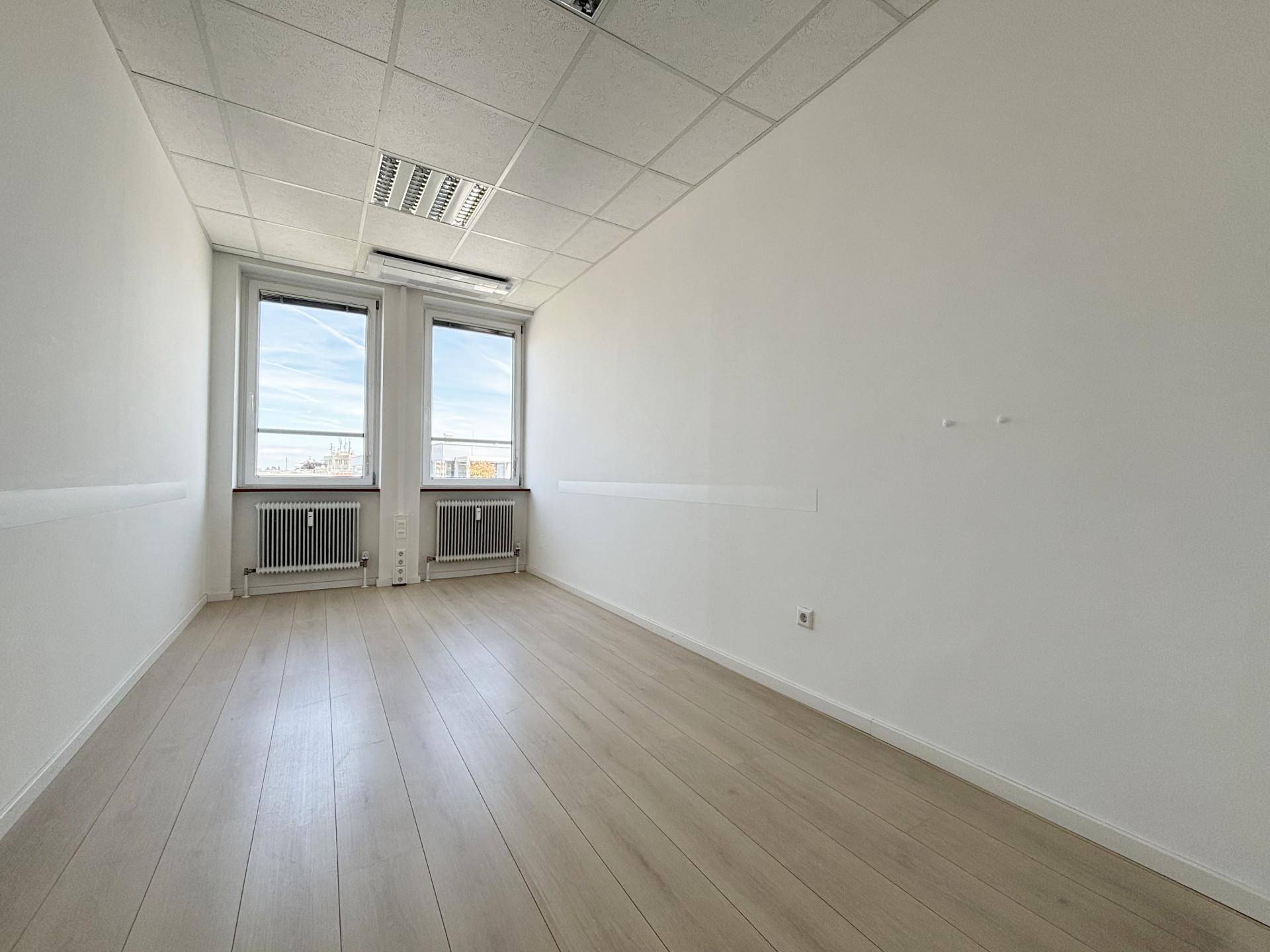 Leeres Büro mit hellem Laminatboden, zwei Fenstern und Heizkörpern unter den Fenstern.