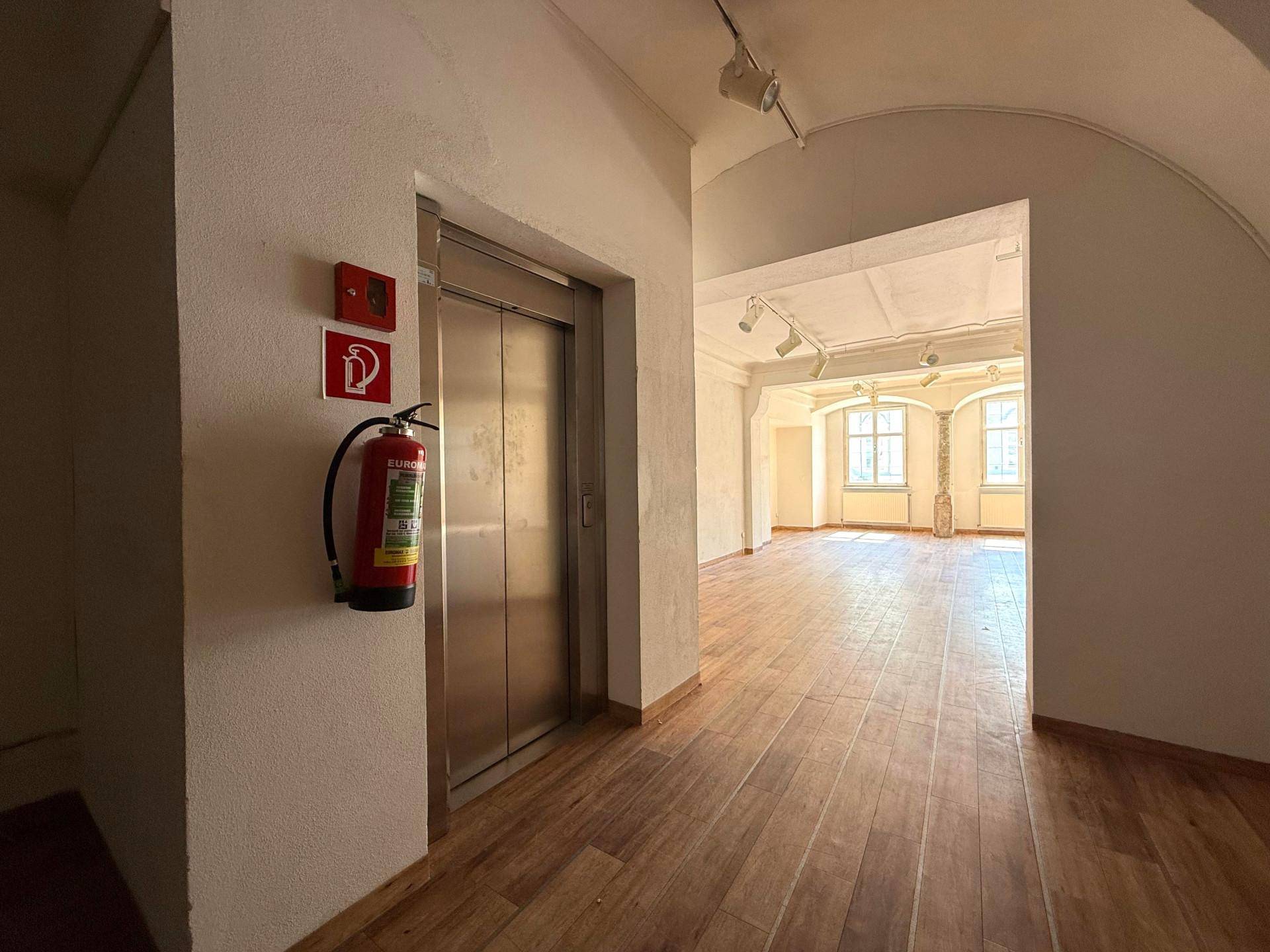 Heller Flur mit Aufzug, Feuerlöscher und Zugang zu weiteren Räumen mit Holzboden und hohen Decken.