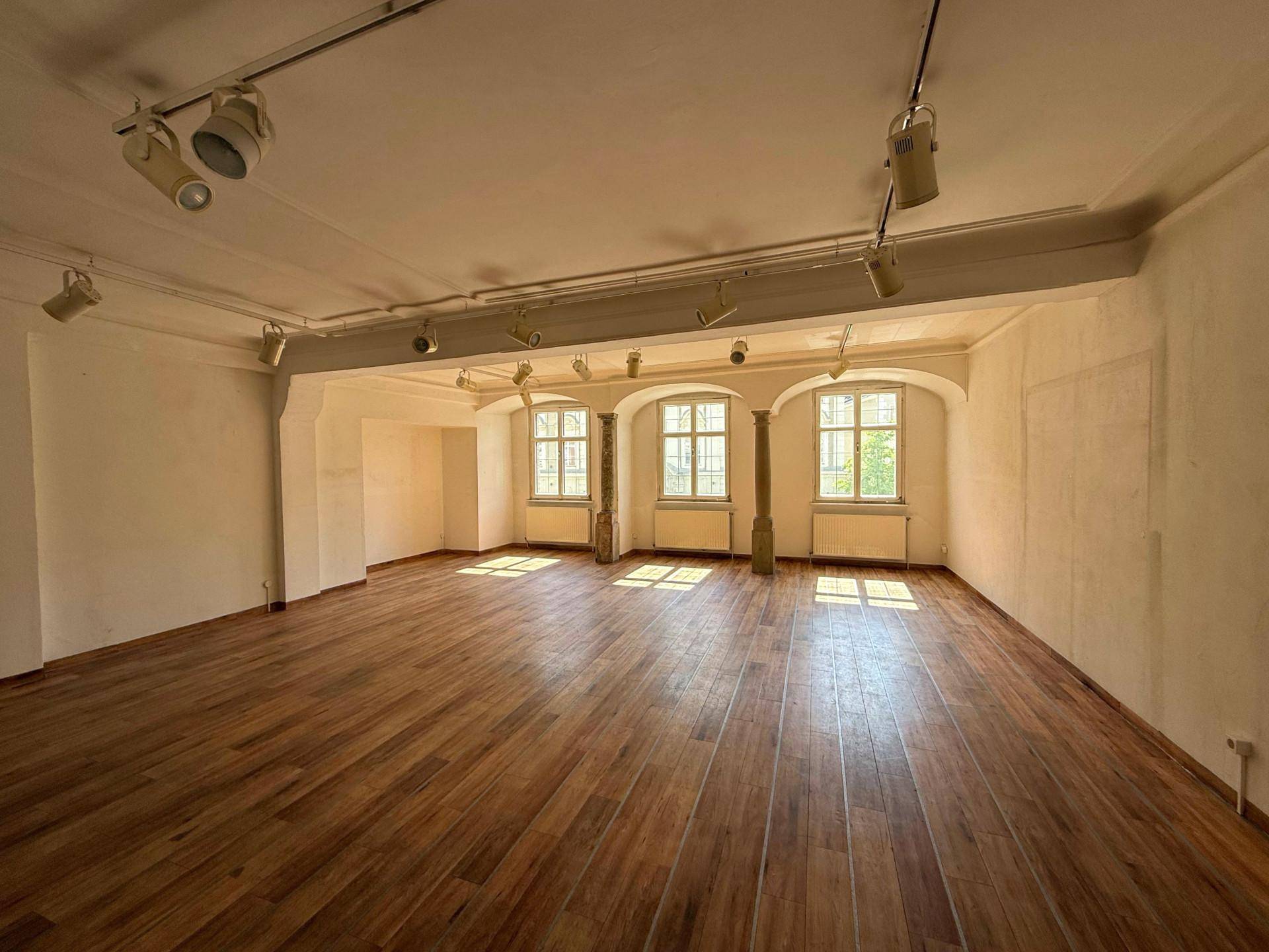 Großer, leerer Raum mit Holzboden, hohen Decken und mehreren Fenstern, die viel Tageslicht hereinlassen.