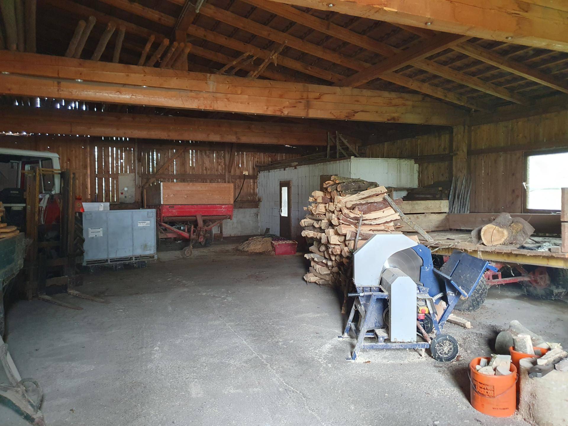 Großer Scheunenraum mit gestapeltem Holz, Sägemaschine und sichtbaren Holzbalken.