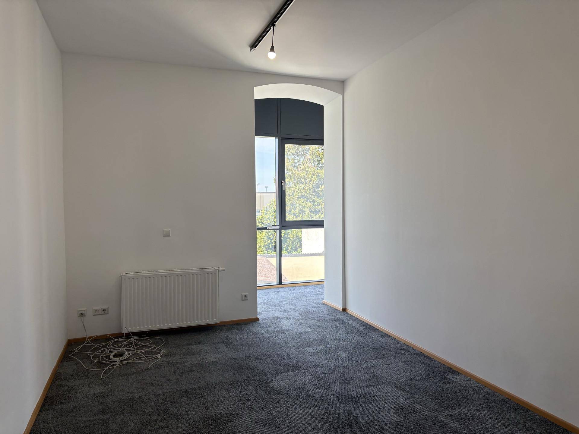 Heller Büroraum mit grauem Teppichboden, weiß gestrichenen Wänden, Heizkörper und einem großen Bogenfenster.