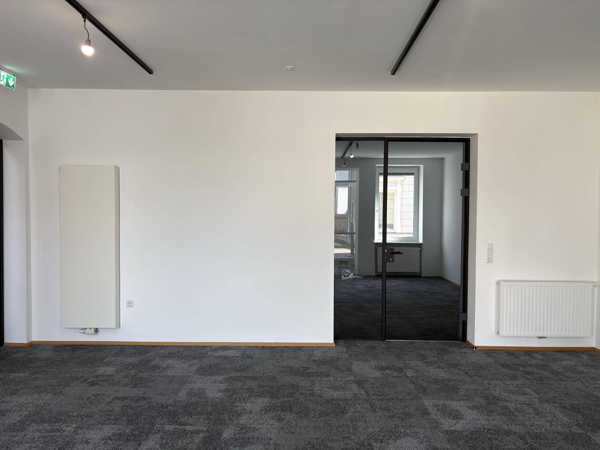 Heller Büroraum mit grauem Teppichboden, zwei Heizkörpern und einer modernen Glastür zu einem Nebenraum.