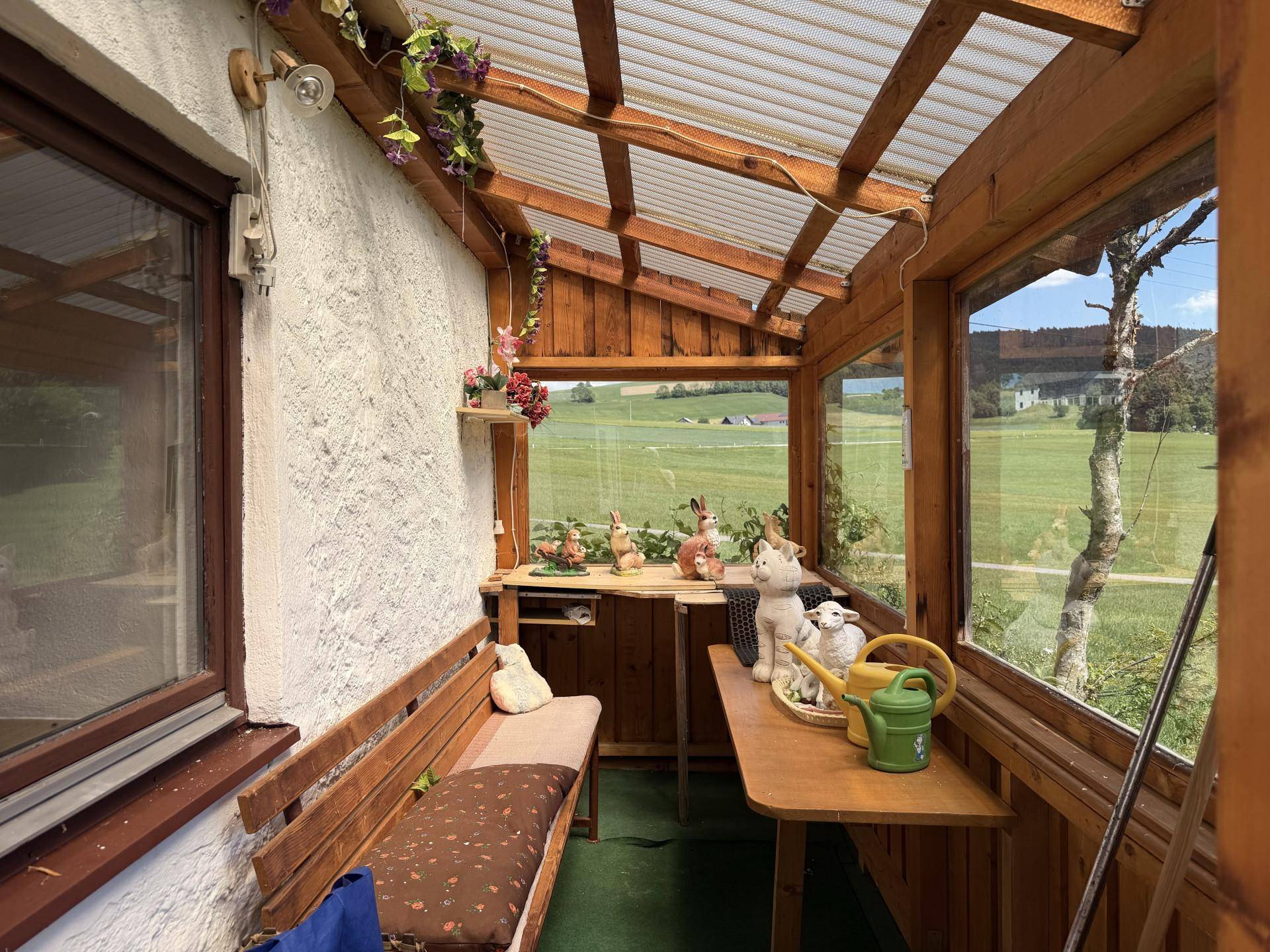 Gemütlicher Wintergarten mit Holzbänken, Dekoration und weitem Blick auf die grüne Landschaft.