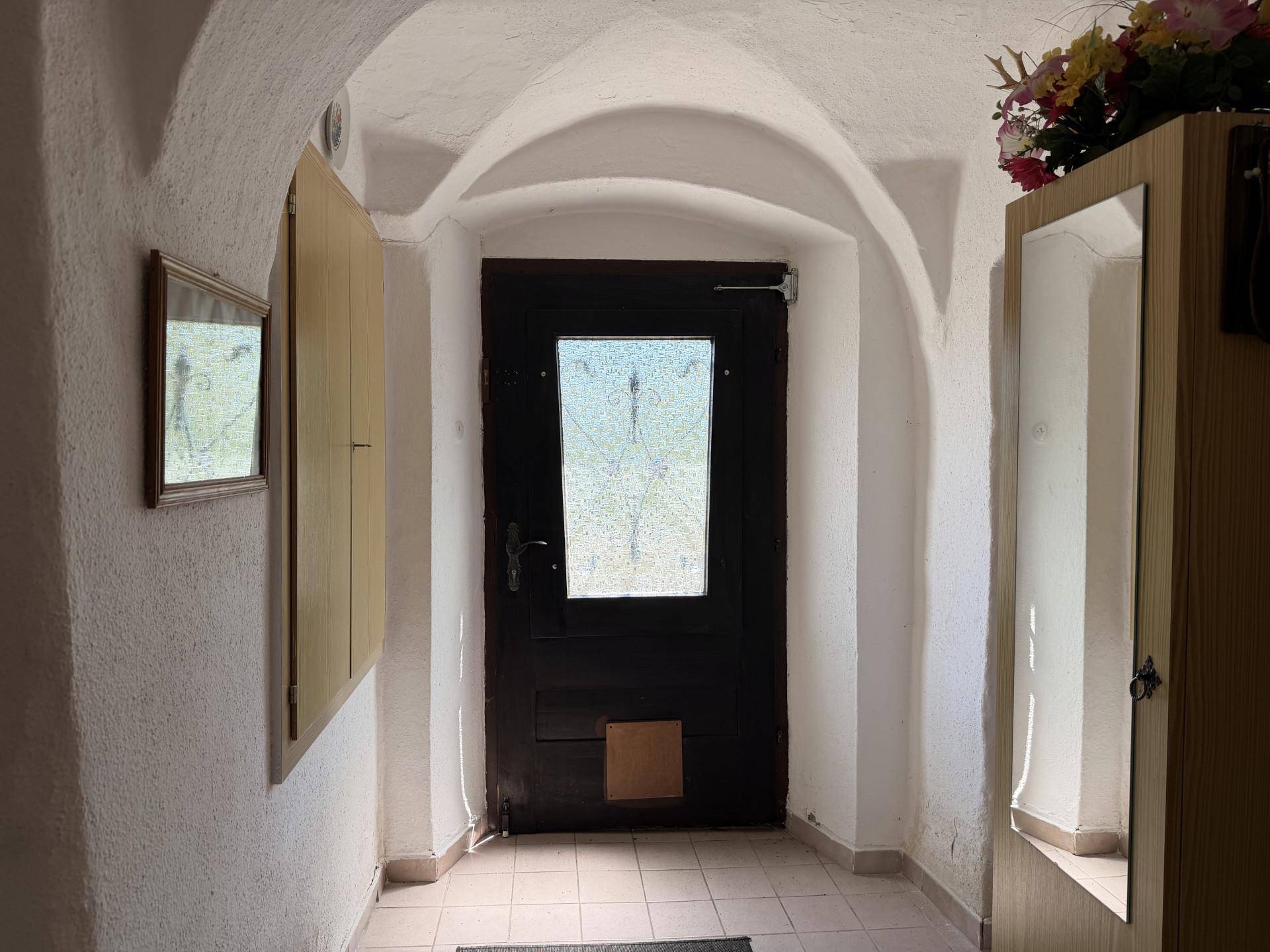Eingangsbereich mit gewölbter Decke, Fliesenboden und Holztür, die zum Haus führt.