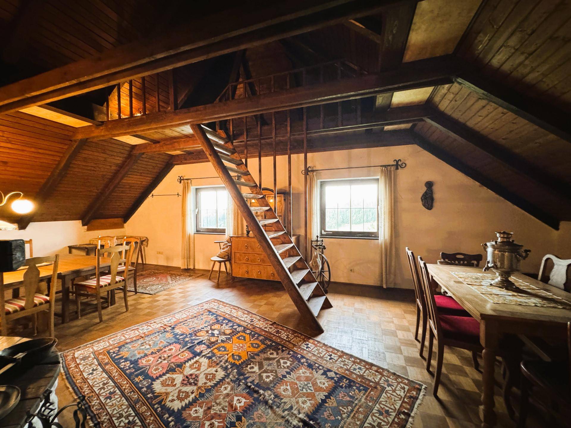 Geräumiger Wohnbereich im Dachgeschoss mit Holzbalken, Teppich und einer Holztreppe zum oberen Bereich.