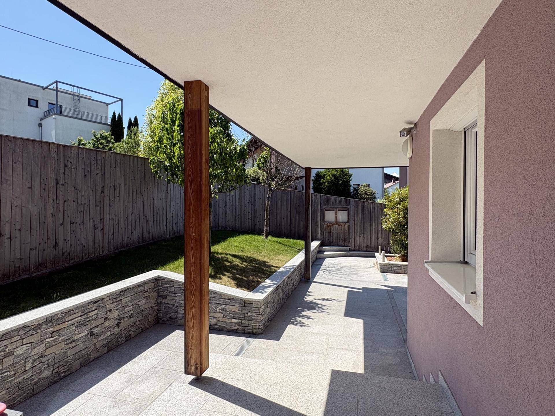 Überdachte Terrasse mit Steinboden, Holzsäule und angrenzender Rasenfläche.