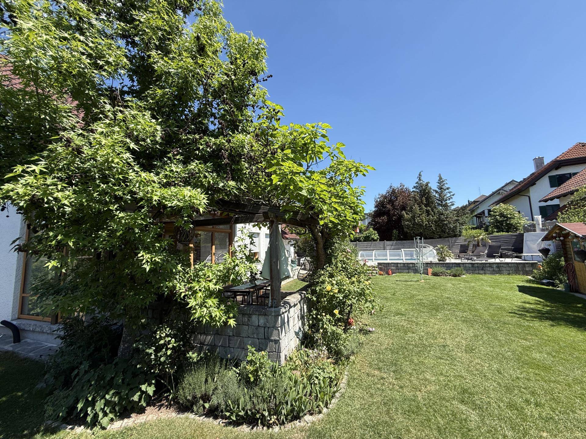 Grüner Gartenbereich mit schattiger Pergola, üppiger Bepflanzung und Pool im Hintergrund.