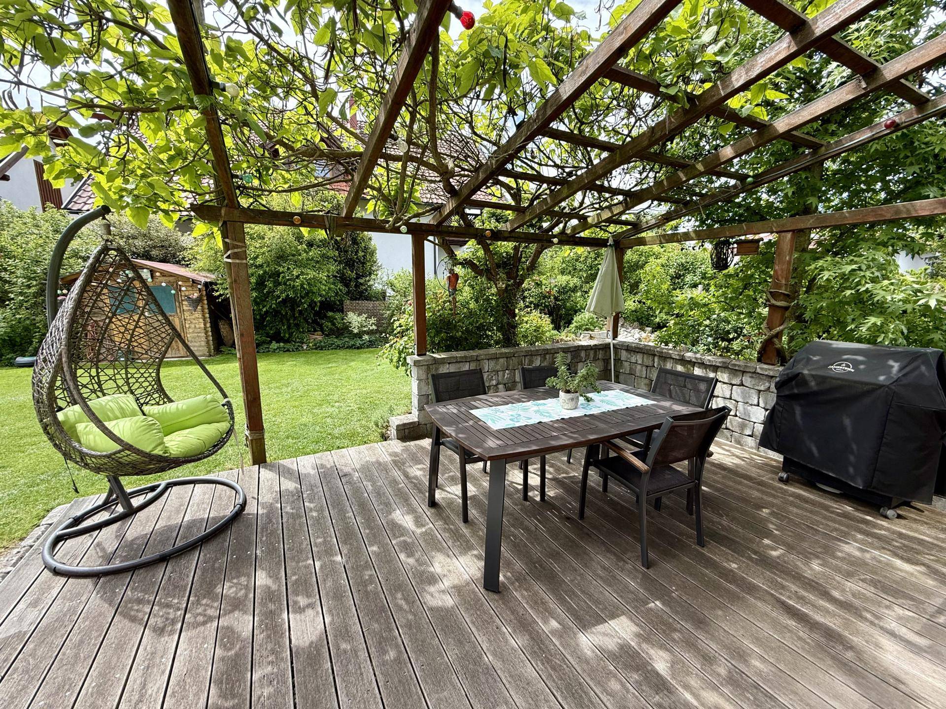 Gemütliche Holzterrasse mit Esstisch, Hängesessel und schattenspendender Pergola im Garten.