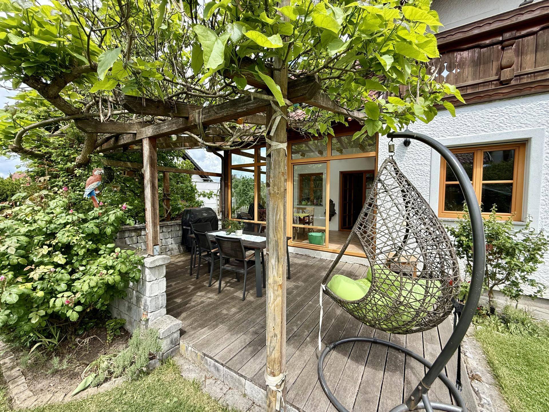 Schattige Holzterrasse mit Esstisch, Hängesessel und üppiger grüner Bepflanzung.
